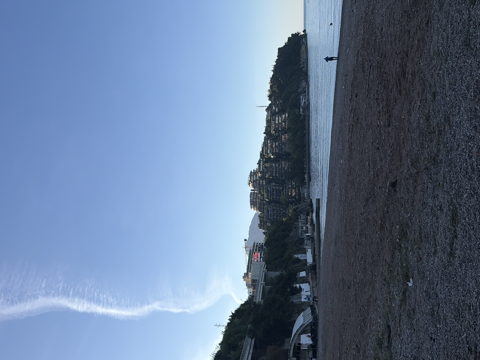 The image depicts a shoreline scene in Budva, Montenegro. A beach occupies the foreground with a gravel surface. Adjacent to the beach is the Adriatic Sea, where a solitary person stands near the water's edge. The coastline is characterized by a steep hill densely populated with multi-story residential buildings. The sky is clear blue with a visible contrail formation. In the background, a mountain is partly obscured by the buildings.