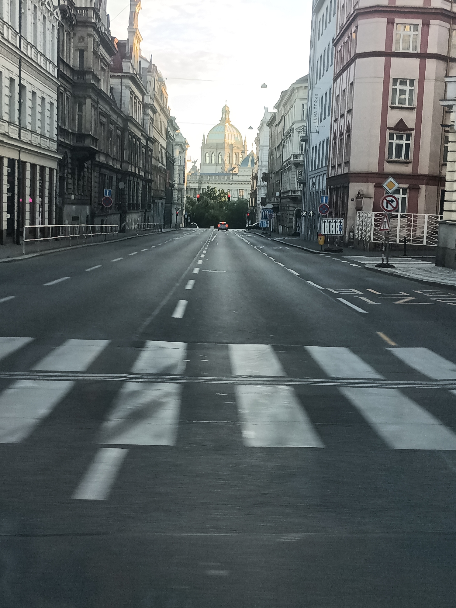 An eye-level, full shot captures a wide, straight road in Prague, Czechia, as viewed from a moving vehicle. The road is lined with old, ornate buildings on both sides, featuring intricate architectural details and varied window styles. In the distance, a large domed building, likely a church or government structure, stands prominently against a bright, overcast sky. Traffic lights are visible, as are various road signs, including pedestrian crossing indicators and directional signs. The road itself is marked with white lines indicating lanes and a clearly defined pedestrian crossing at the forefront of the image, which is blurred due to motion. A few cars are visible further down the road, and trees are interspersed along the sides. The overall impression is one of an urban street scene in a historic European city.