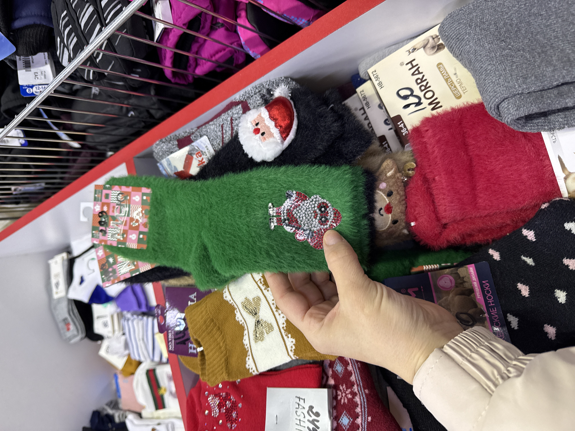 Here is a description of the image:
The image is taken in what appears to be a store in Beloyarsky, Russia. The focus is a shelf filled with various socks. There are Christmas-themed socks in the selection, including designs with Santa Claus, reindeer, and holiday decorations. A person's hand is visible, holding a green, fuzzy sock with a sparkly Christmas ornament. The socks are neatly stacked and displayed in a red-edged shelving unit. There are also packages of socks visible, some with Russian text printed on them, indicating product information. Shelves are present above the socks with clothes.