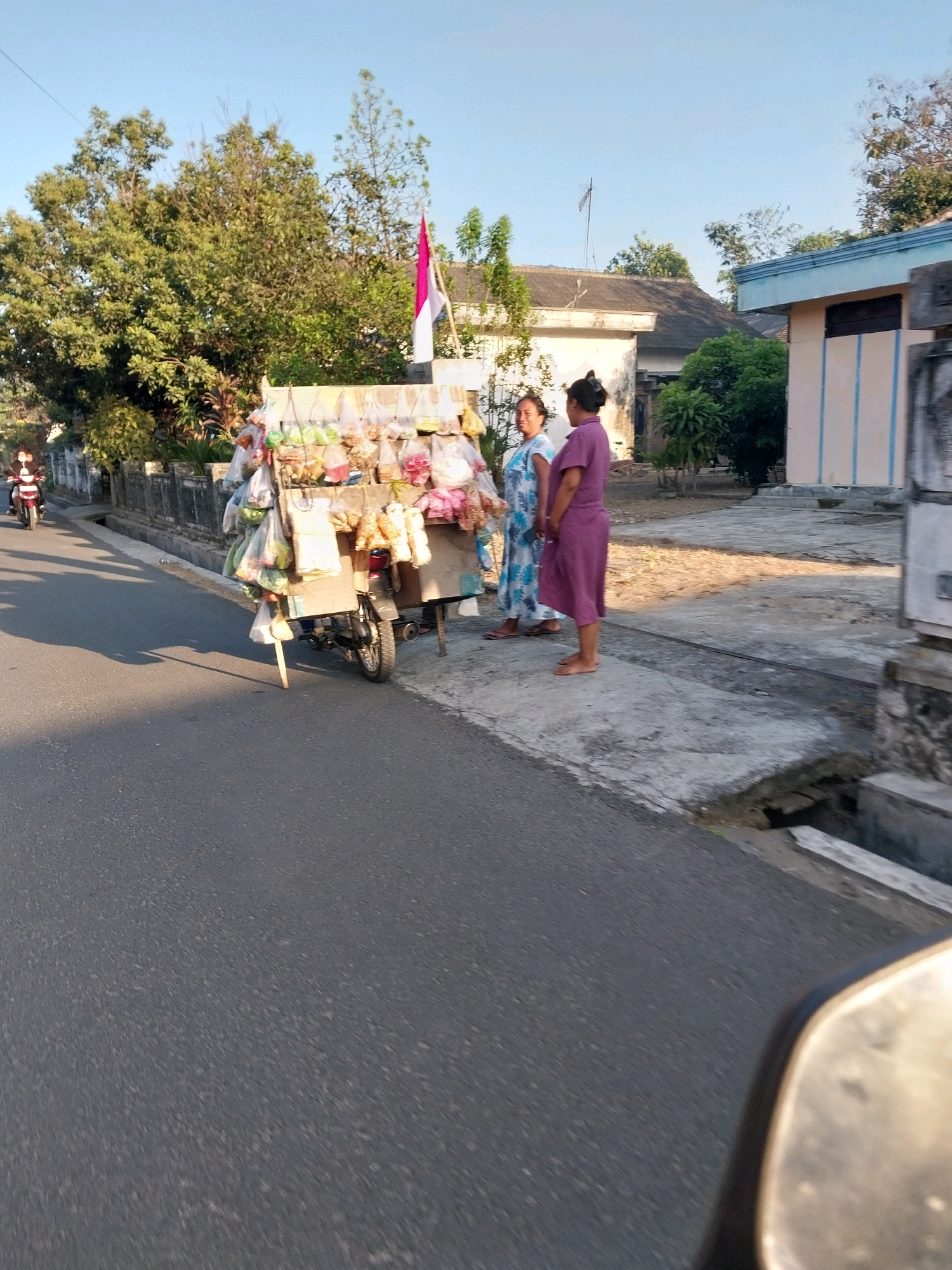 In the city of Nglegok, Indonesia, a street scene unfolds under a clear sky. A motorbike, likely used for a mobile cart, is parked on the side of a paved road, a common sight for street vendors. The cart itself is laden with numerous plastic bags filled with various goods, likely snacks or small items for sale. Two women stand beside the cart, appearing to be engaged in conversation or attending to customers. One woman wears a blue and white patterned dress, while the other is in a purple dress. Behind them, a building with a light-colored facade and blue trim is visible, along with lush green trees and vegetation. The presence of a red and white flag, possibly the Indonesian flag, suggests a local or national context. The overall scene depicts a typical day of commerce and community life on a street in Nglegok.