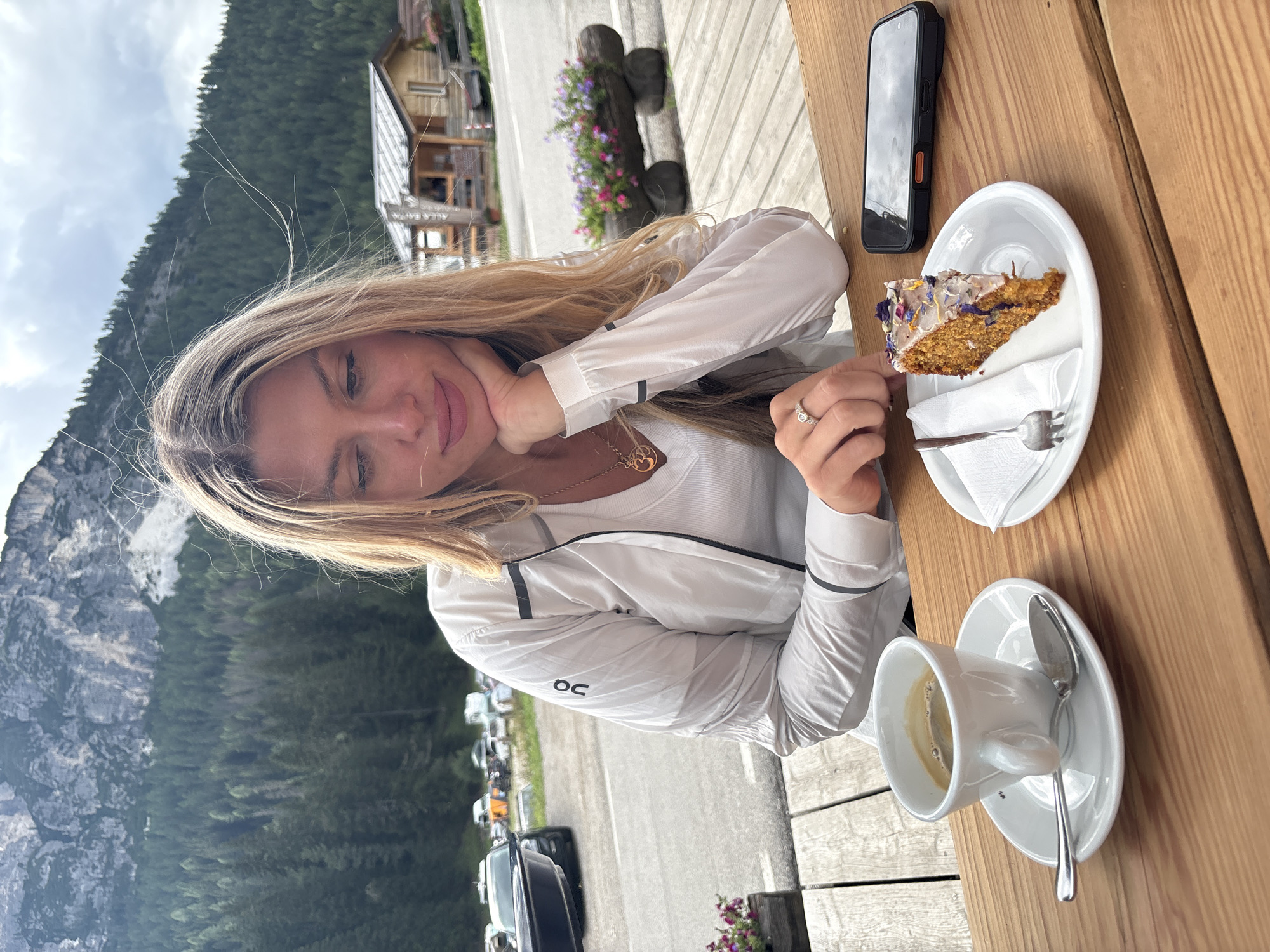 A young Caucasian woman with blonde hair is seated at a wooden table outdoors. She is wearing a white jacket over a white top and a necklace. Her left hand is resting on her chin, and she is wearing a ring on her left index finger. In front of her on the table are a cup of coffee with a saucer and spoon, a slice of cake on a plate with a fork and napkin, and a smartphone. The background shows a mountainous landscape with pine trees, a wooden building, and parked cars. The scene appears to be at a cafe or restaurant in Auronzo di Cadore, Italy.