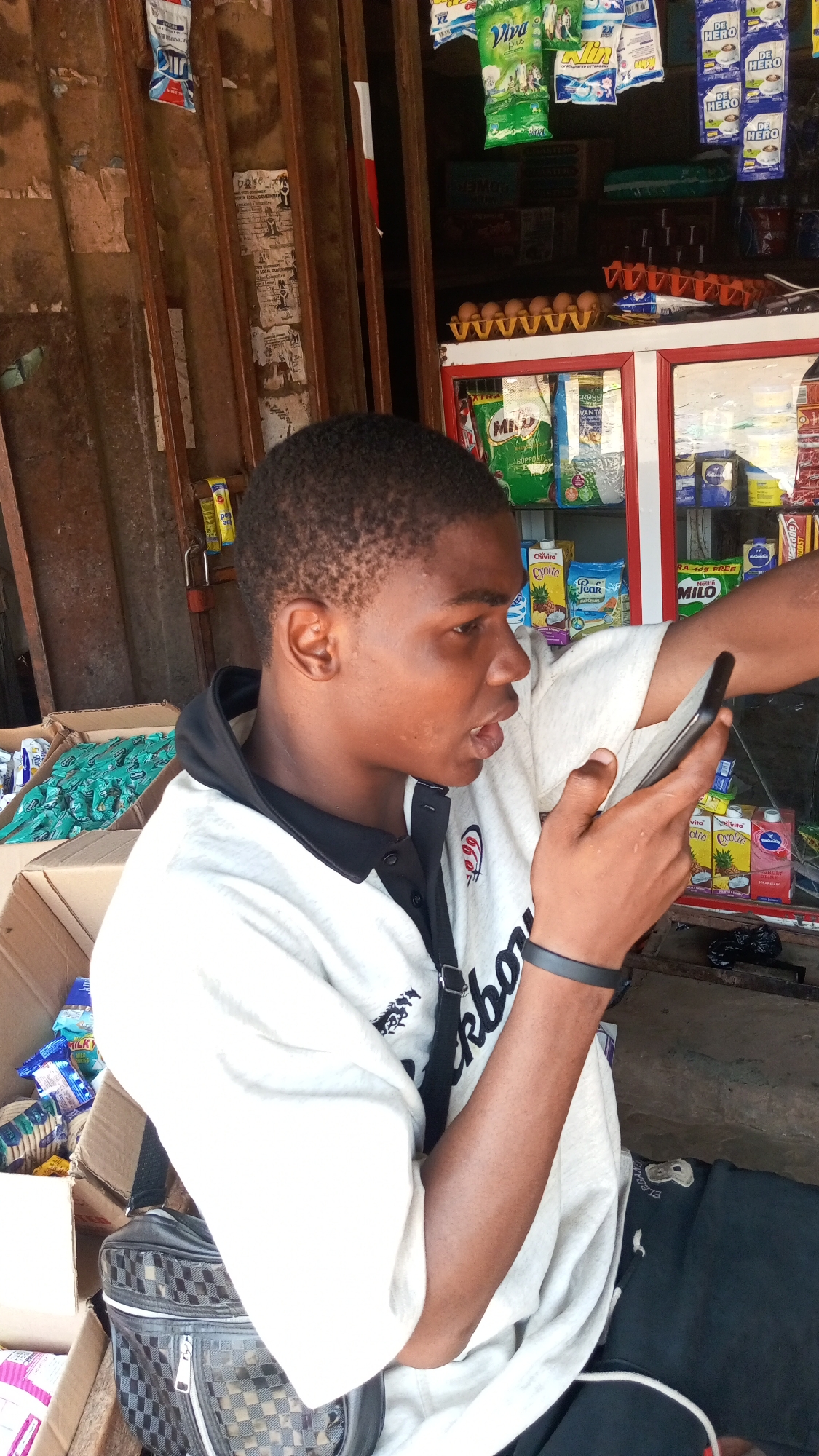 The image shows a young man with short, dark hair looking at a smartphone in his hand, his expression suggesting he is speaking or otherwise engaged with the device. He is wearing a white t-shirt with black trim and lettering on the side. A black strap crosses his shoulder. He is seated beside a black and silver shoulder bag.
The background indicates he is near a small store or kiosk. The store features a display case stocked with various food and beverage products, including juice boxes, milk cartons, and packages of Milo and other food items. Cartons of eggs are visible on top of the display case. There are also several advertisements and packages hanging from the store's structure.
The overall setting appears to be Onitsha, Nigeria, based on the products and the style of the store. The image is taken in daylight and seems to be captured at eye level.