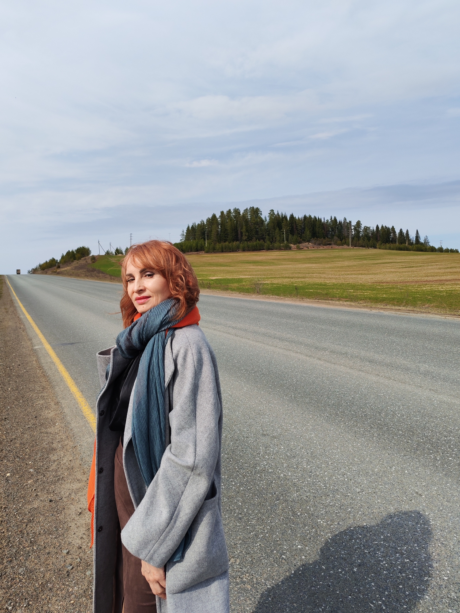 A woman stands on the shoulder of a two-lane highway in Нечкино, Russia. She has red hair and is wearing a gray coat, a blue scarf, and an orange undershirt. To the right of the road is a field with a hill and trees in the background. To the left of the road is a grassy shoulder. The sky is cloudy.
