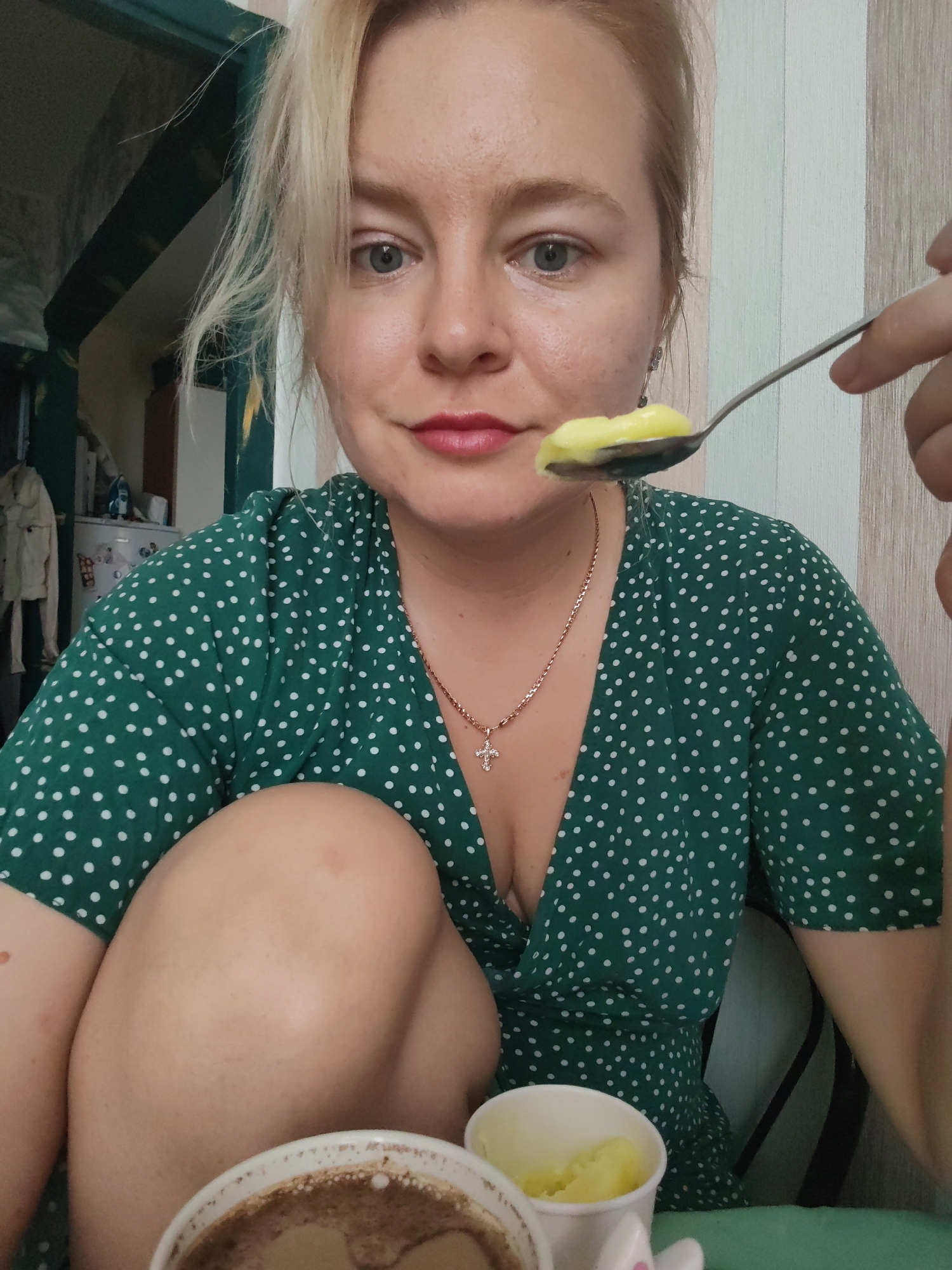 In Izhevsk, Russia, a woman with blonde hair is captured in a close-up shot, holding a spoon with a portion of yellow food towards her mouth. She is wearing a green dress with white polka dots and a necklace with a cross pendant. In the foreground, a white cup with a brown beverage and another white cup with yellow food are visible. The background shows a wall with vertical stripes and some furniture.