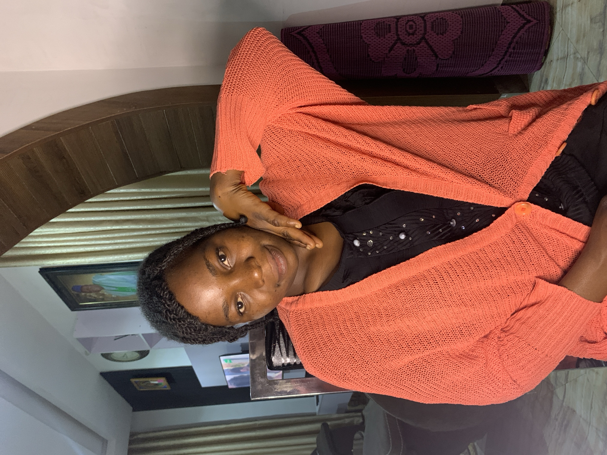 The image shows a Black woman with braided hair, wearing a black top with decorative beads and an open coral-colored knitted cardigan. She is seated and looking towards the camera, with her right hand touching her neck and chin. The background suggests an indoor environment, possibly a living room in Jos, Nigeria, with architectural details like an arched wooden frame, curtains, framed pictures on the wall, and a clock. A rolled-up mat with a floral pattern is visible on the right side of the frame. The lighting appears to be artificial, creating shadows and highlighting the subject.