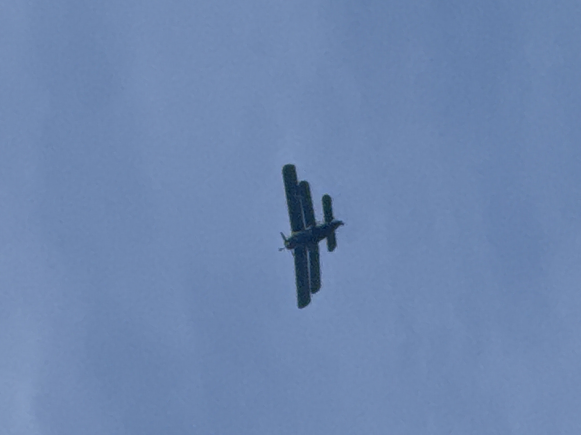 The image displays a dark-colored biplane aircraft in flight against a uniform light blue sky. The aircraft, observed from a low angle and slightly ahead, is oriented with its nose pointing towards the upper-left quadrant of the frame. It features a biplane wing configuration, with two main wings stacked vertically, and a propeller assembly is visible at the front. The sky is clear and cloudless. No other subjects, objects, or environmental details are discernible. The location is Unknown, Unknown.