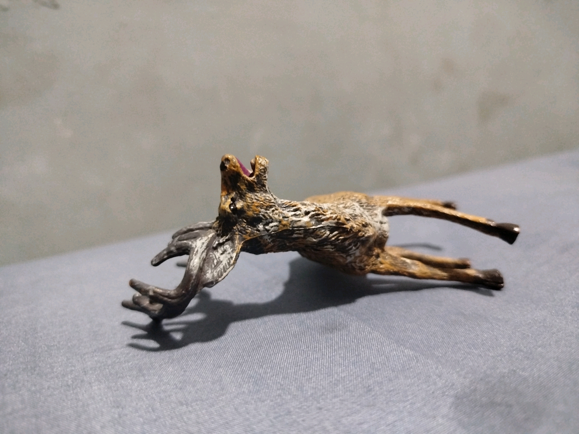 A static shot captures a detailed, small figurine of a deer in mid-leap. The figurine, made of a material that appears to be resin or a similar composite, is predominantly brown with white and black accents highlighting its fur texture. Its mouth is open, revealing a pink tongue, and its head is tilted upwards, suggesting a dramatic pose. The deer's antlers are dark brown and branch out intricately. The figurine is positioned horizontally across a textured, gray surface, possibly a fabric cloth, which is illuminated from the side, casting a distinct shadow to the left of the figurine. In the background, a plain, textured gray wall serves as the backdrop, devoid of any features. The image does not depict any specific urban or natural setting within Mojoagung, Indonesia.