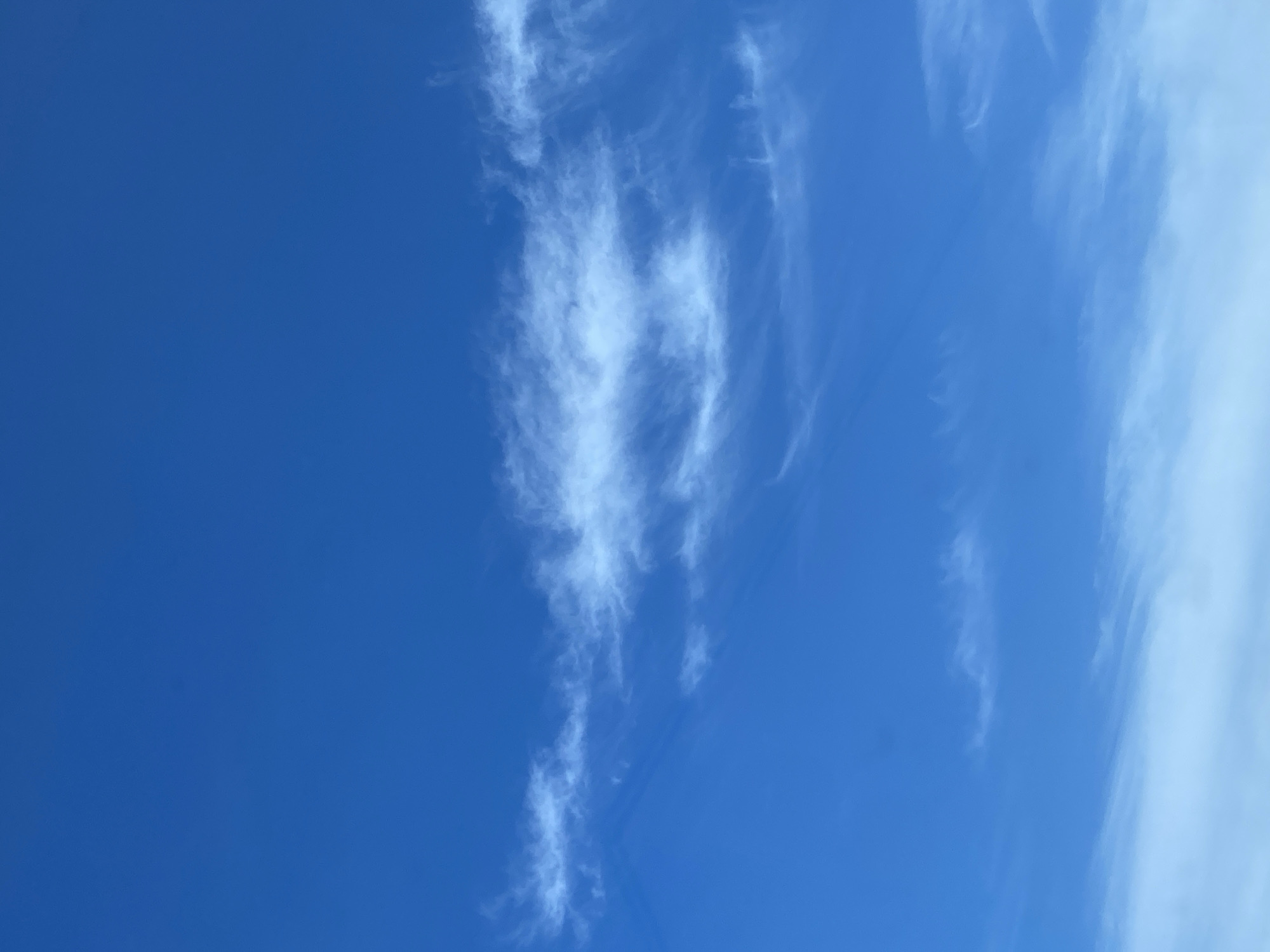 The image shows a clear blue sky with thin, wispy white clouds. The clouds are scattered across the upper half of the frame, with a prominent band of clouds running horizontally across the middle. The bottom portion of the image is dominated by a lighter, more diffused layer of clouds that fades into the blue of the sky. There are no people, subjects, or objects visible in the image other than the sky and clouds. The scene is consistent with an outdoor environment, likely during the daytime.