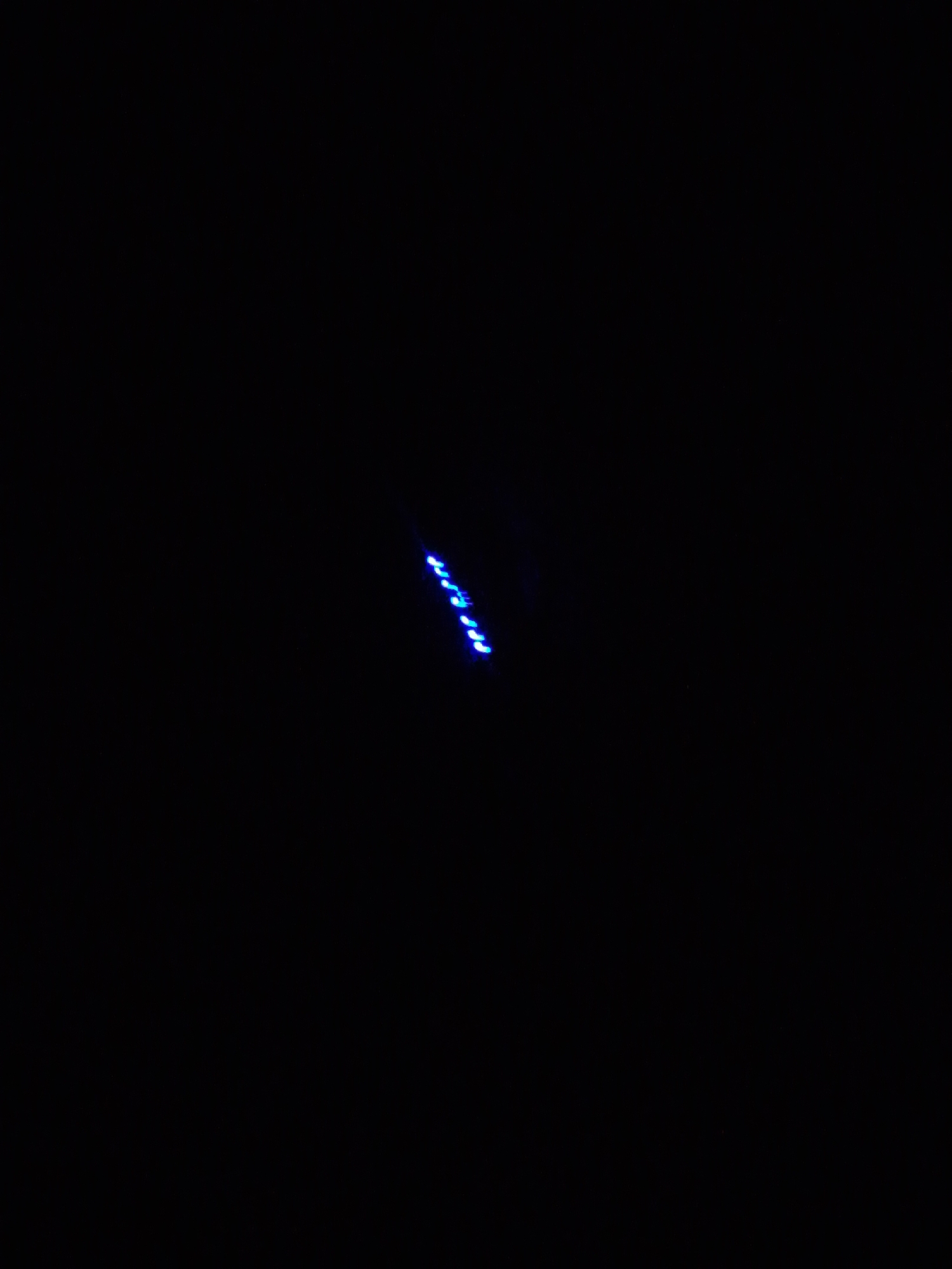 The image depicts a nocturnal or extremely low-light scene in the city of Keffi, Nigeria. The frame is dominated by an entirely black background, indicating a complete absence of ambient illumination. The primary subject is a diagonal formation of approximately twelve distinct, intensely bright blue light sources. These light points originate in the upper-right quadrant and extend towards the lower-central portion of the frame, forming a slightly curved, broken line. The specific objects or devices emitting these blue lights are not discernible. No individuals, additional objects, or environmental features are visible within the scene due to the pervasive darkness.
