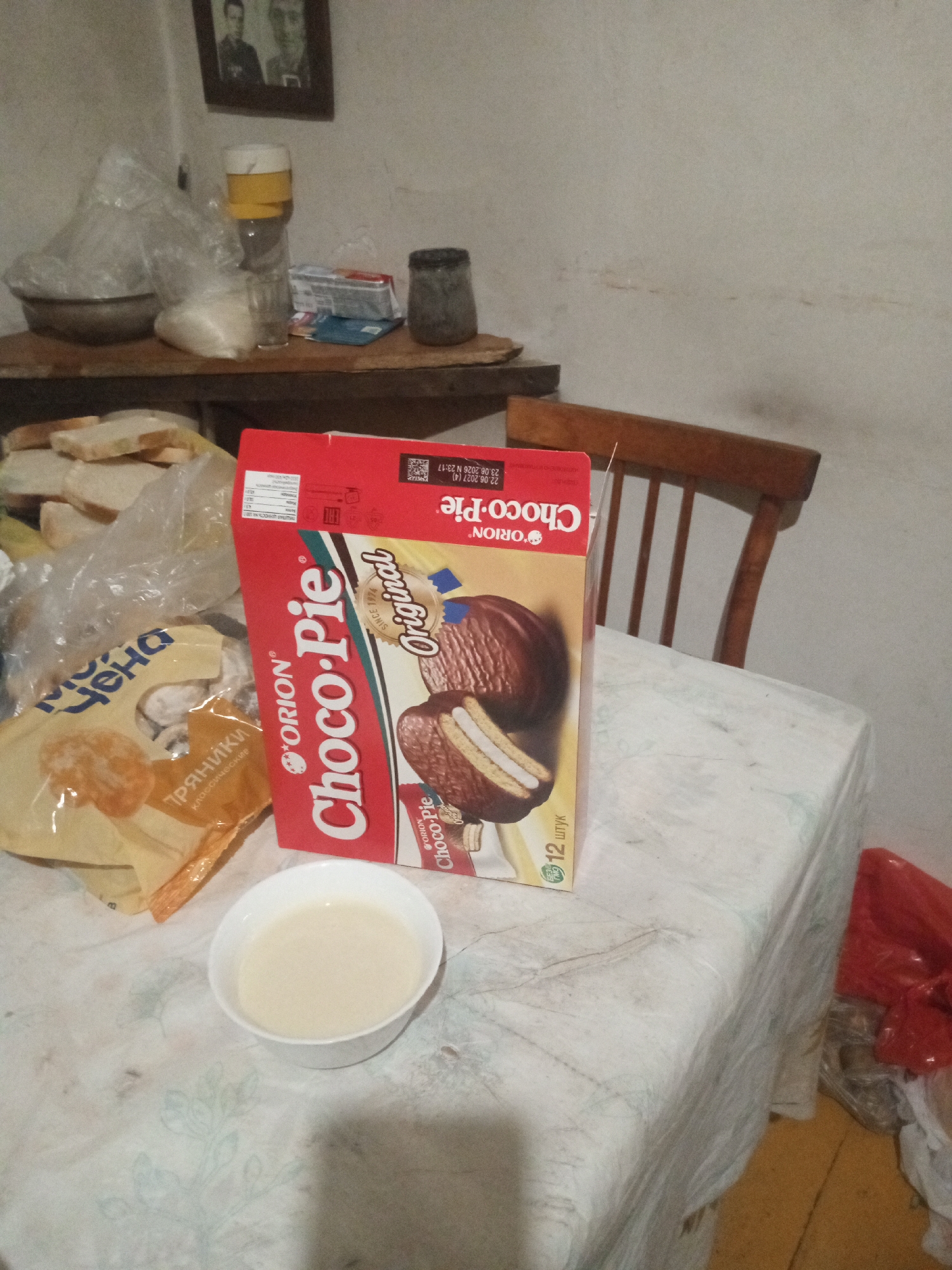 The image depicts a table set with food items, possibly in a home kitchen in Rubtsovsk, Russia. A box of Orion Choco-Pie cookies is prominently displayed on the table, next to a small white bowl filled with a light-colored liquid, likely milk or cream. To the left of the Choco-Pie box, a yellow plastic bag, also containing food items, is partially visible. In the background, on a shelf, are various household items including a glass jar with a yellow lid, a bag of white powder, and a dark, cylindrical object. A wooden chair is positioned behind the table. The overall impression is one of a simple, domestic setting.