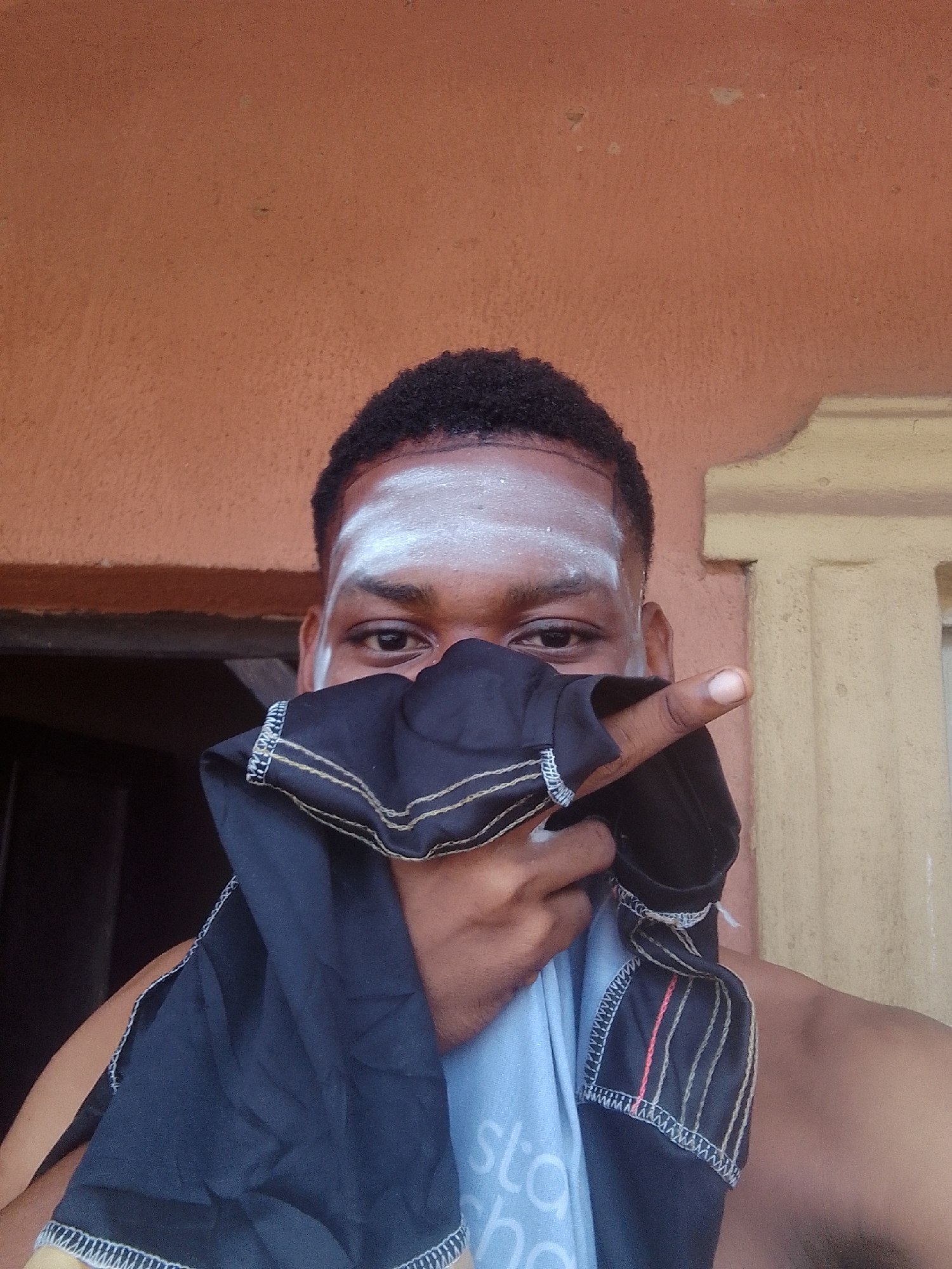 A close-up, waist-up, color photograph depicts an individual with dark skin, short curly black hair, and a white powdery substance on their forehead. The person is positioned outdoors, with a peach-colored wall behind them. The subject covers the lower part of their face with a dark blue cloth trimmed with white, gold, and red stitching. They are pointing their right index finger towards the right side of the frame. Part of the torso, shoulders, and a light blue shirt with the word "sta" visible are in the frame. The location appears to be in Nkpor, Nigeria, based on contextual information not visible in the image.