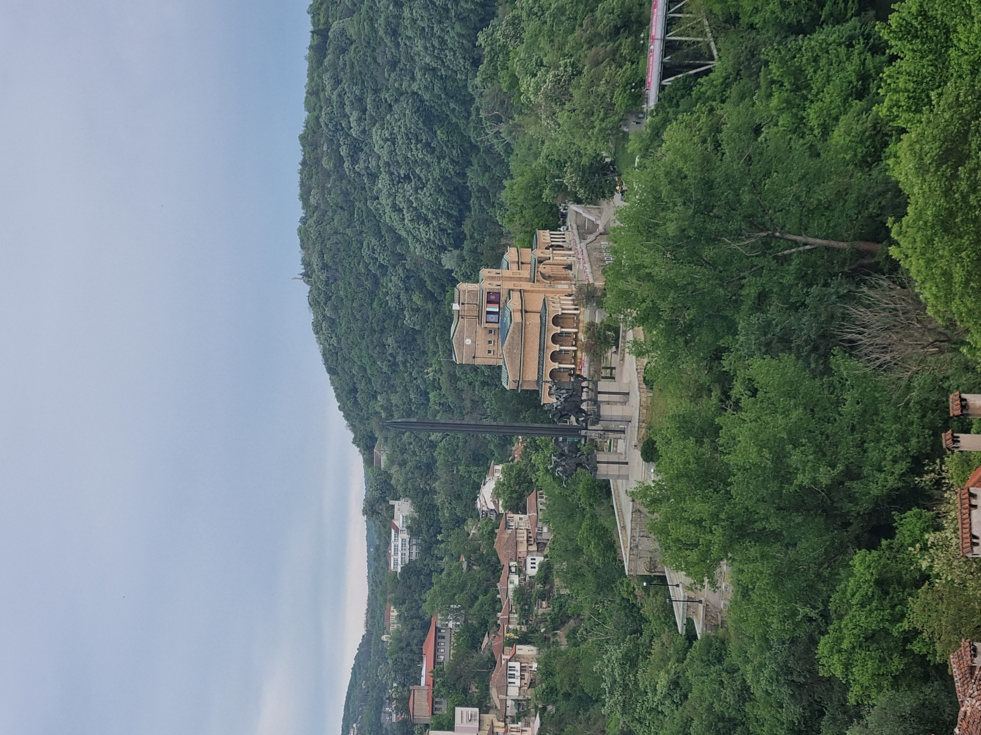 The image shows a view of Veliko Tarnovo, Bulgaria, featuring the Monument to the Asen Dynasty. The monument is a prominent structure with equestrian statues and a tall, sword-like obelisk. Behind the monument stands a building with arched windows and a clock. The foreground is filled with lush green trees. In the background, there is a hillside covered in dense forest, as well as residential buildings with red-tiled roofs nestled among the trees. The sky is overcast.