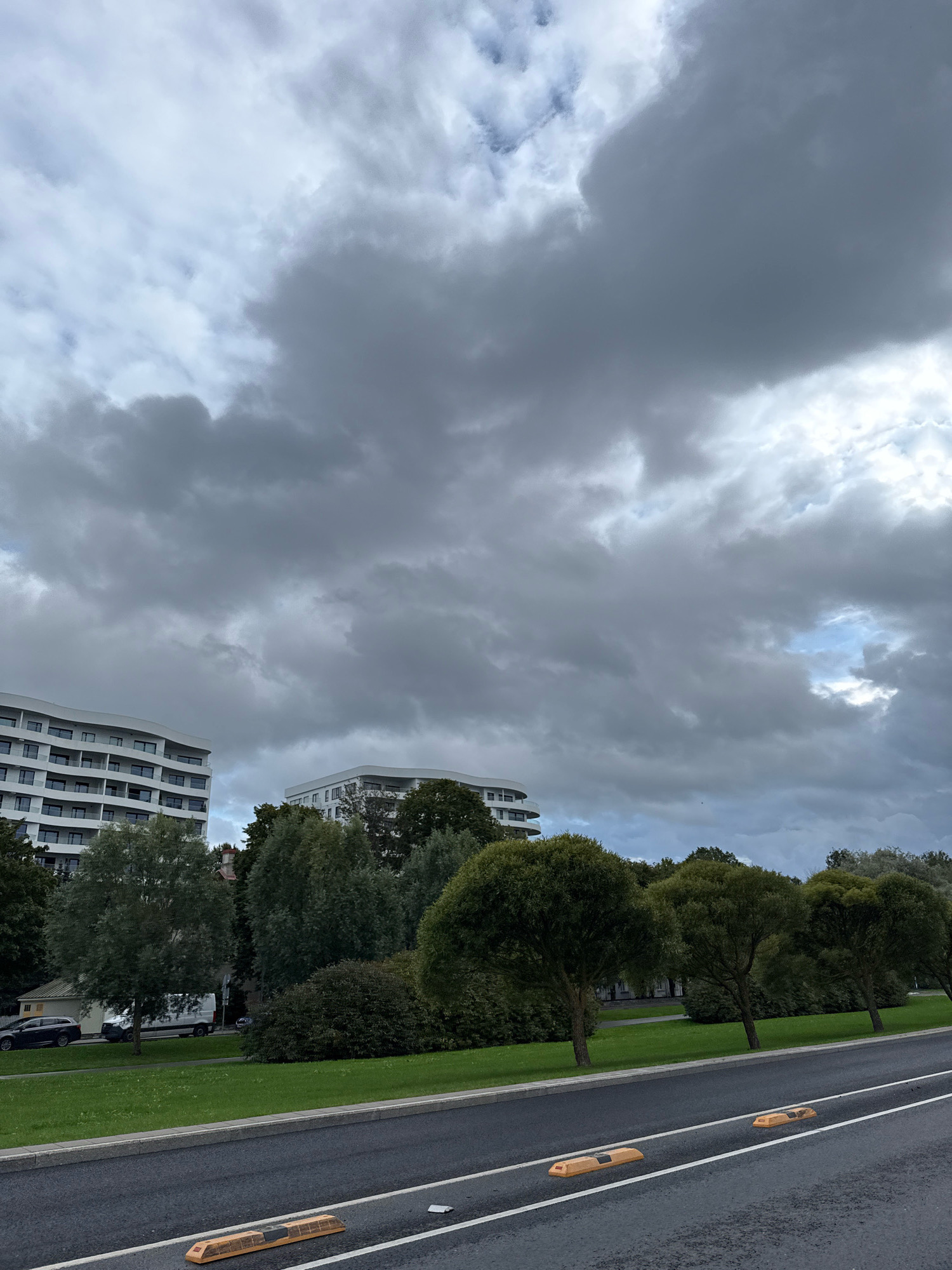 This is a medium-distance, eye-level, horizontal, full-color photograph of a street scene in Tallinn, Estonia. The sky is heavily overcast with dark, dramatic clouds, but a small patch of blue sky is visible to the upper right. In the background, two modern, white, multi-story buildings with numerous windows are visible, partially obscured by lush green trees. The trees in the foreground are rounded and dense. The foreground consists of a paved road with yellow parking guides and white lane markings. A grassy verge runs alongside the road, separating it from the trees and buildings. Several cars are parked on the side of the road in the middle ground. The overall impression is of a slightly gloomy day in an urban environment with abundant greenery.