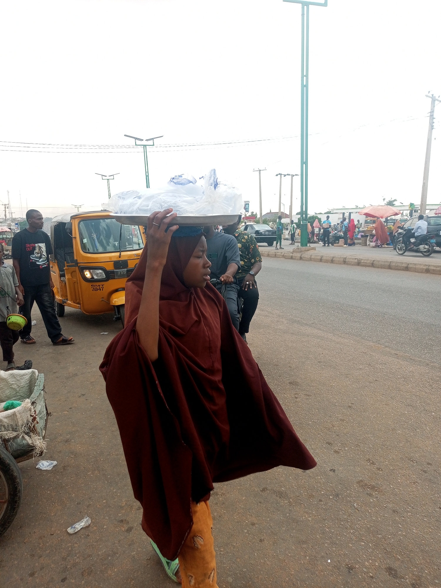 A medium-shot captures a woman in the foreground, adorned in a maroon hijab and robe, balancing a tray laden with wrapped items on her head. Her arm is raised to steady the tray. The background features a bustling city street in Unknown, Unknown. A yellow three-wheeled vehicle, a man in a "Skate Out" T-shirt, and a boy with a green bowl stand near her. The street is lined with people, vehicles, and market stalls. A power line is visible in the sky.