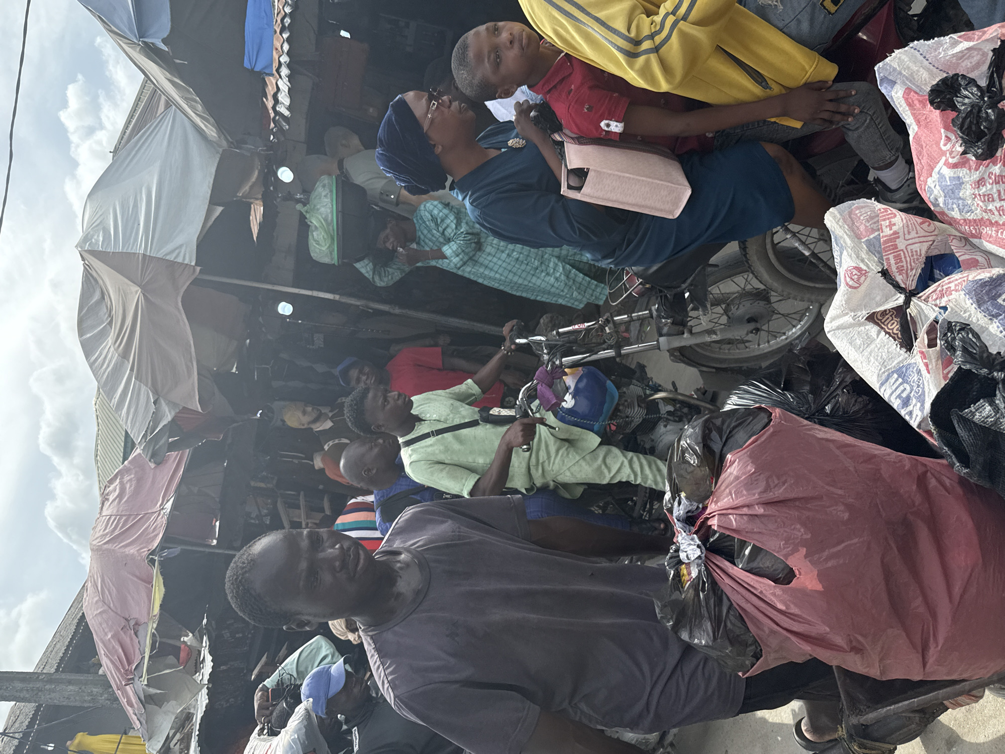This image captures a vibrant, crowded outdoor market or street scene, consistent with urban environments in Ado, Nigeria. The scene is brightly lit, indicating daytime with a visible sky and scattered white clouds overhead.
The foreground is dominated by a man in a dark grey t-shirt, who looks towards the viewer while holding a red plastic bag. To his right, several large, woven plastic bags, some white with red/blue text such as "PURE SUGAR" and "CHOCO," are stacked, tied shut with black plastic.
In the mid-ground, a woman in a blue headwrap (gele) and blue dress, wearing glasses and carrying a light pink handbag, looks towards the left of the frame. Beside her, a young boy in a red polo shirt and blue jeans looks directly at the camera. Behind them, a man in a light green long-sleeved shirt is seated on a motorcycle, holding the handlebars, with a blue helmet resting nearby. Other individuals are visible throughout the frame, including a person in a light blue patterned shirt and others, contributing to the sense of a bustling crowd.
The setting features makeshift market stalls or shelters constructed from various fabrics and tarpaulins stretched over metal frames, providing shade. Clothes or textiles are seen hanging from these overhead structures. The ground is largely obscured by people and goods, reinforcing the crowded nature of the location. The overall atmosphere is one of lively commerce and daily activity.