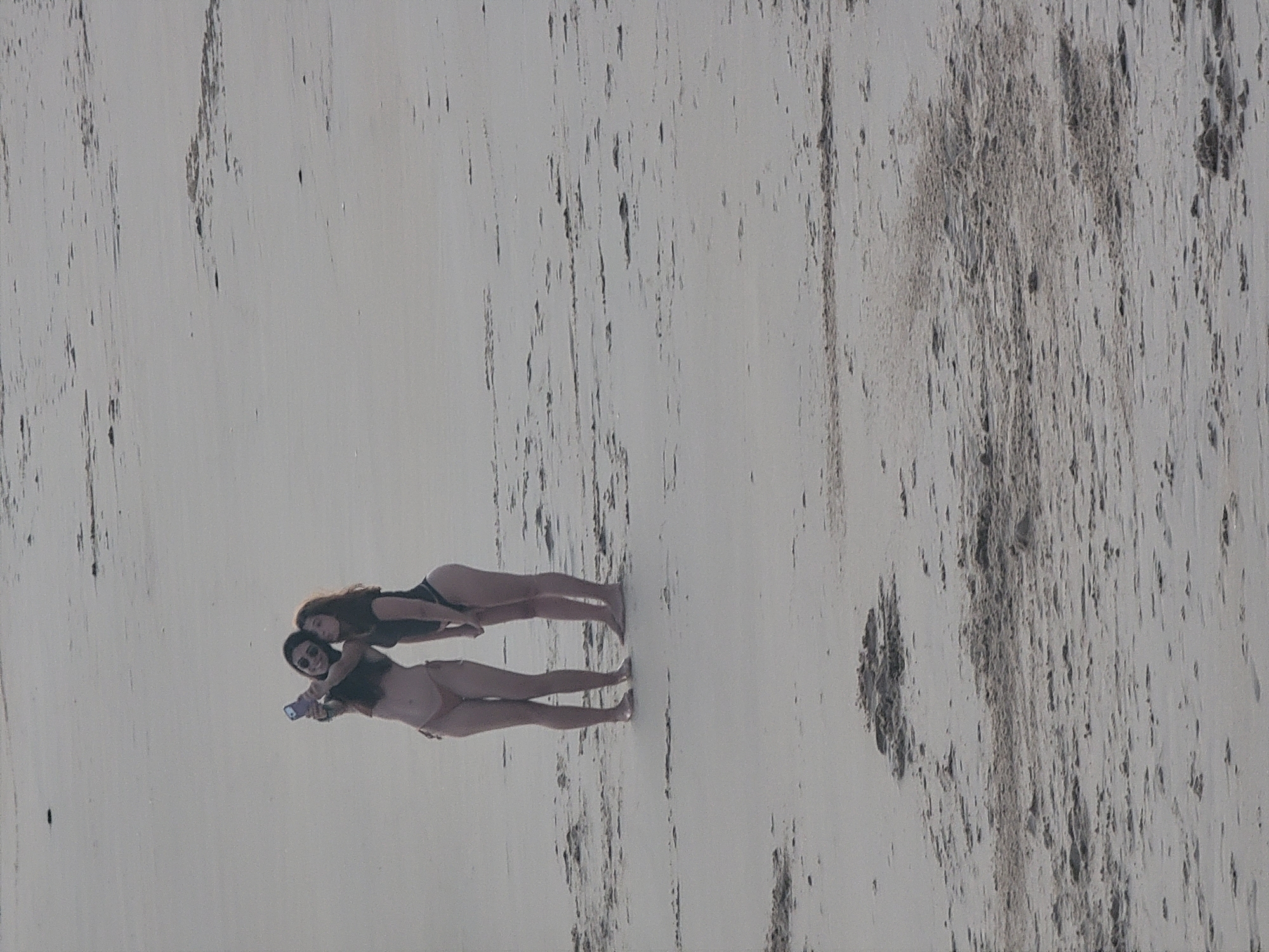 Two young women are standing on a sandy beach, taking a selfie with a smartphone. The woman on the left is wearing a bikini and holding the phone. The woman on the right is also wearing a bikini. They are both facing the camera. The background is a sandy beach with some ripples in the sand. The scene is consistent with a location on the coast of Quarteira, Portugal.