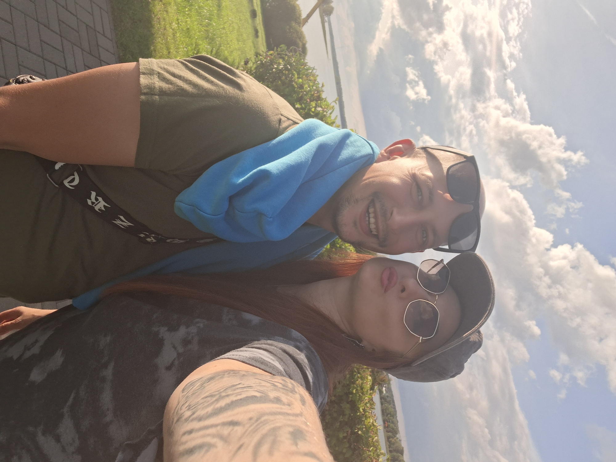 A selfie is taken from a low angle outdoors, with a man on the left and a woman on the right. The man is wearing a green t-shirt, a blue scarf around his neck, and sunglasses. He is smiling and looking at the camera. The woman is wearing a dark gray t-shirt and a baseball cap, and her lips are pursed as if she is kissing the camera. She is also wearing sunglasses. Her right arm, which is holding the camera, is visible in the foreground, showing a tattoo. Behind them, there is a body of water, likely a lake, surrounded by greenery and trees. The sky is cloudy, with some sunlight breaking through. The foreground shows a paved walkway and grass. The scene is located in the city of Myadzyel, Belarus.