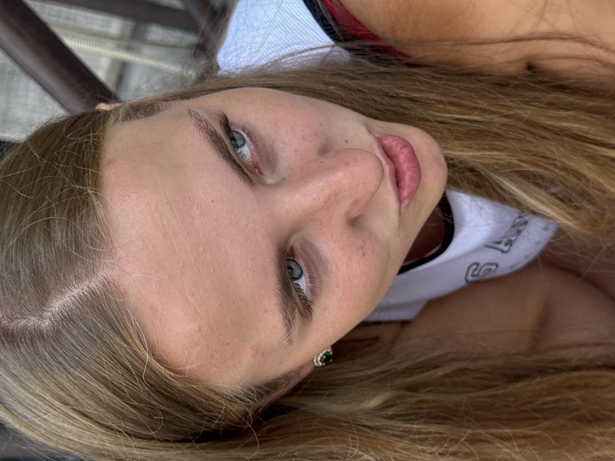 The image is a close-up, high-angle shot of a young woman with blonde hair, looking upwards and to the right. She is wearing a white tank top with black trim and a dark-colored garment underneath. A green earring with a dark stone is visible in her left ear. The background appears to be an outdoor setting with blurred wooden structures, suggesting a patio or seating area. The lighting indicates it is daytime. The specific location in Trieste, Italy, cannot be determined from the visual information provided.