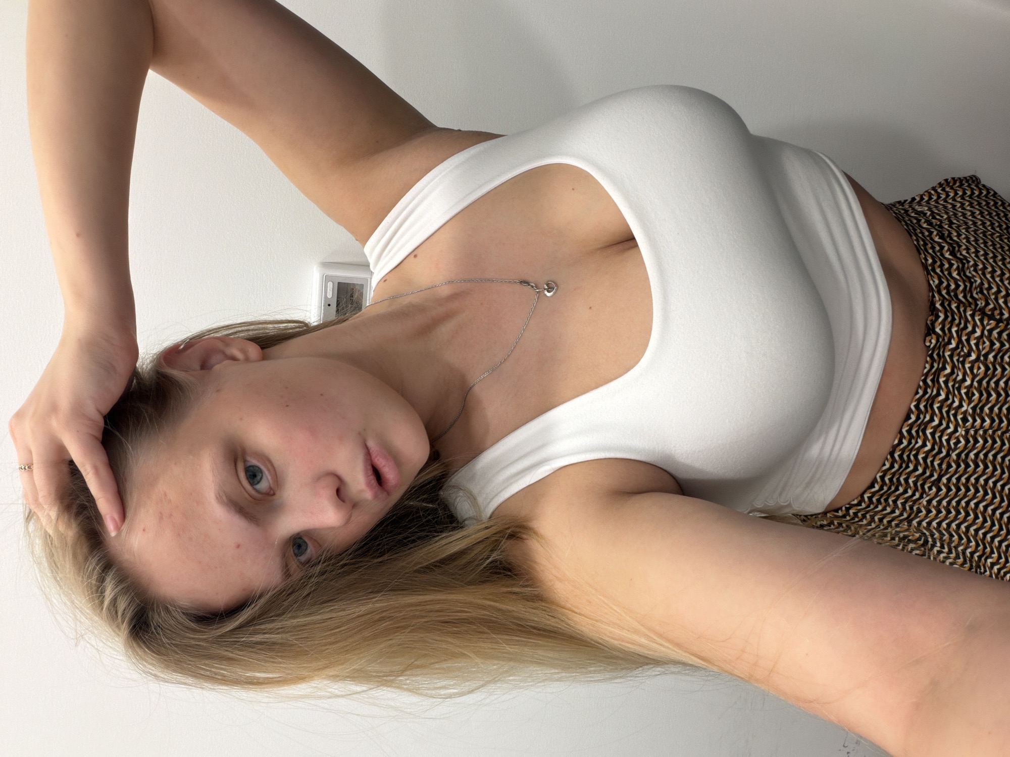 The image displays a young woman with long, straight blonde hair and light blue eyes, captured in a close-up shot from an upward angle. She is lying down, with her head slightly tilted to the left, looking directly at the camera. Her right arm is raised, with her hand gently resting near her hair.
She is wearing a snug-fitting white tank top or crop top that reveals her midriff. A delicate silver chain necklace with a small, heart-shaped pendant adorns her neck and collarbone. Her lower body is clad in shorts or loose pants featuring a distinctive brown, black, and white geometric zig-zag pattern.
The background consists of a plain, unadorned white wall, suggesting an indoor setting. A white rectangular object, possibly a thermostat or an electrical control panel, is mounted on the wall behind her head. The lighting is soft and even, without strong shadows, indicating a well-lit indoor environment. Her skin is fair, with some minor freckles or blemishes visible on her forehead. There is no clearly discernible text in the image. The scene takes place in Izhevsk, Russia.