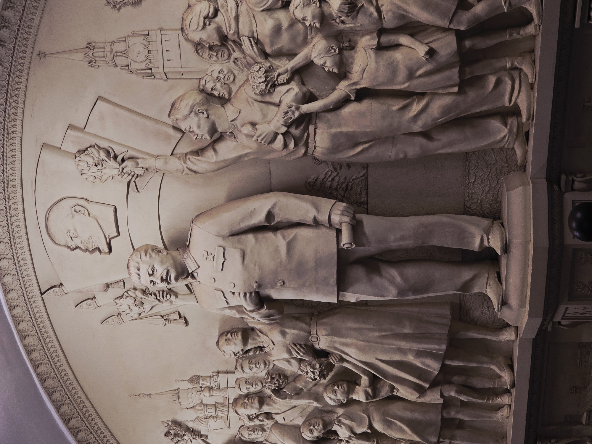 A bas-relief sculpture is depicted in the image. It features a full-body depiction of Vladimir Lenin. The top and bottom edges of the work show people holding flowers and buildings, as well as people holding flowers and buildings. The sculpture is located in an unknown city in an unknown location.