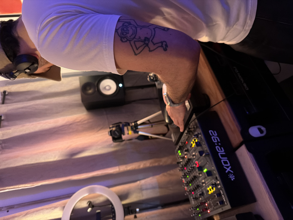 A man with a monkey tattoo on his arm stands in what appears to be a home studio setup. He wears a white T-shirt, cap, and headphones. On a wooden desk, there's a Xone:92 DJ mixer with illuminated controls. A studio monitor speaker is visible next to a camera mounted on a tripod. A ring light stands near the bottom of the image, and the walls are draped with fabric. A Denon DJ device is positioned behind the mixer.