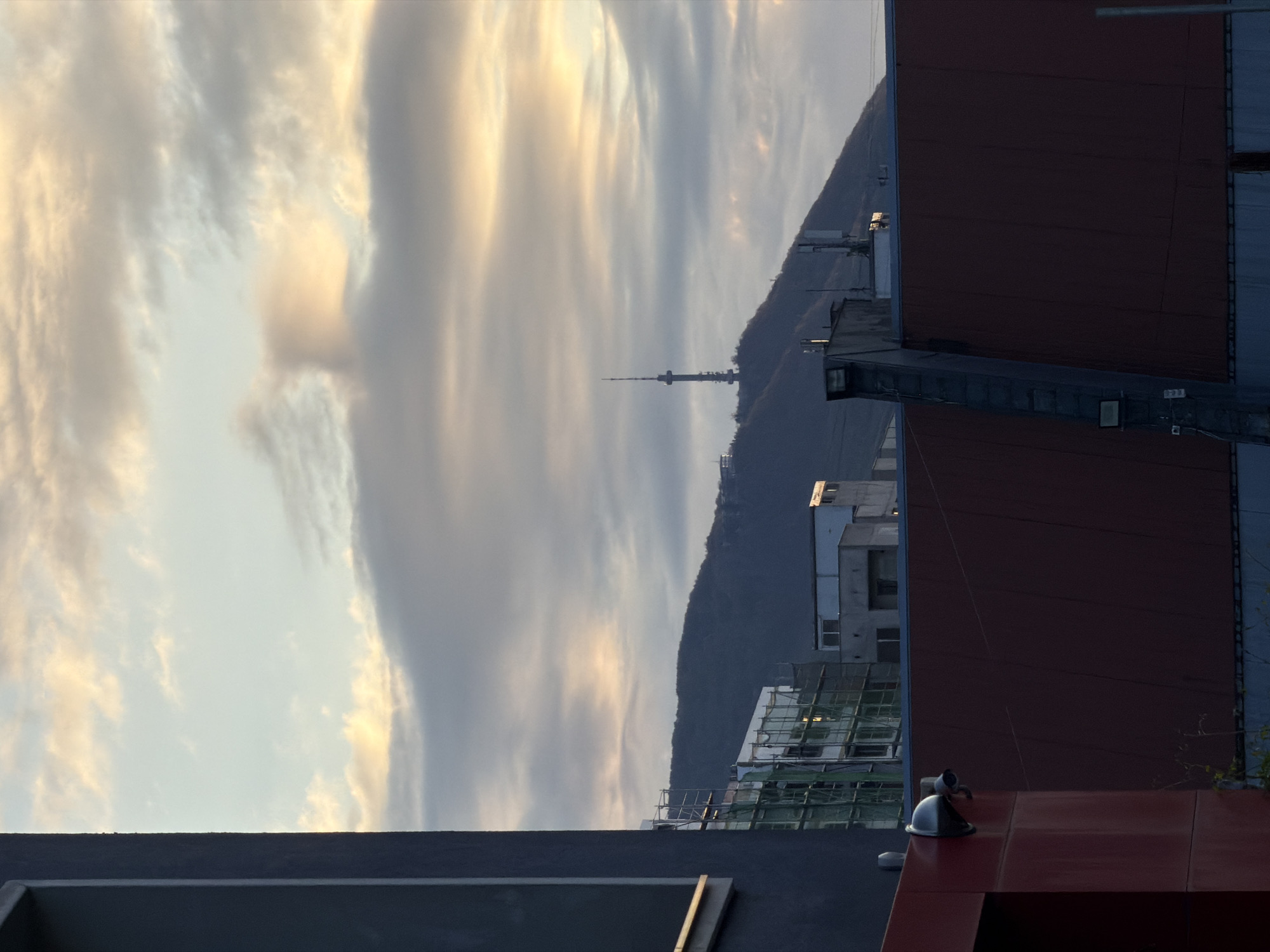 The image depicts an urban scene with a view of a hillside and cloudy sky. In the background, a television tower rises from the hill, which likely represents the Kopitoto area on Vitosha Mountain in Sofia, Bulgaria. The foreground features sections of buildings with varying heights and construction stages, including modern architecture and scaffolding. A person is seated on a rooftop, near a surveillance camera. The overall scene evokes a sense of urban landscape juxtaposed against the natural environment.