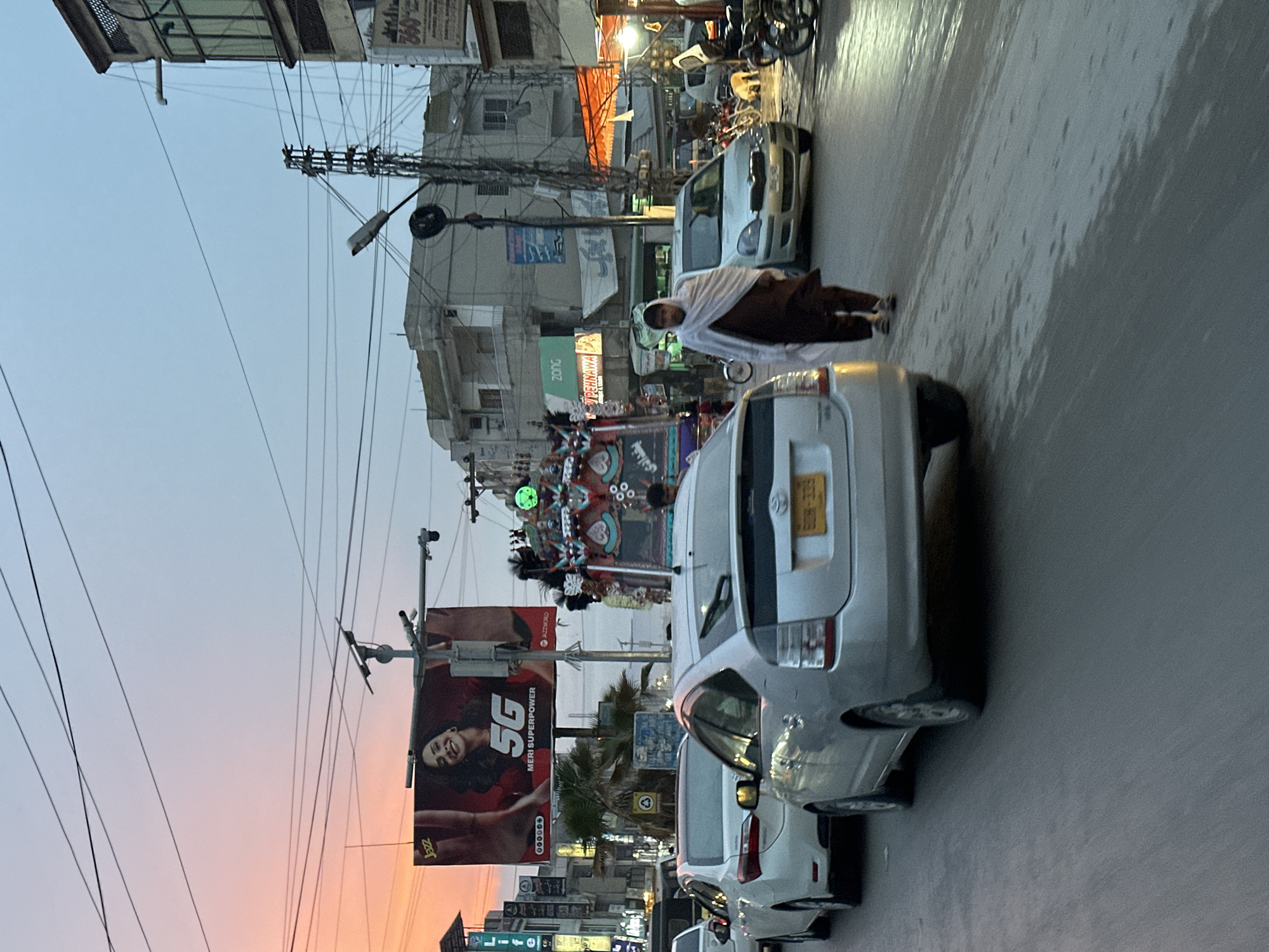 The image depicts a street scene in Quetta City Tehsil, Pakistan. The sky shows an orange-to-blue gradient, suggesting either sunrise or sunset. The street is lined with buildings and various businesses. A large billboard on the left side promotes 5G internet services. Several cars are parked or moving on the right side of the street. A man wearing traditional Pakistani clothing walks beside the cars. Overhead, there are numerous electrical wires and light poles.