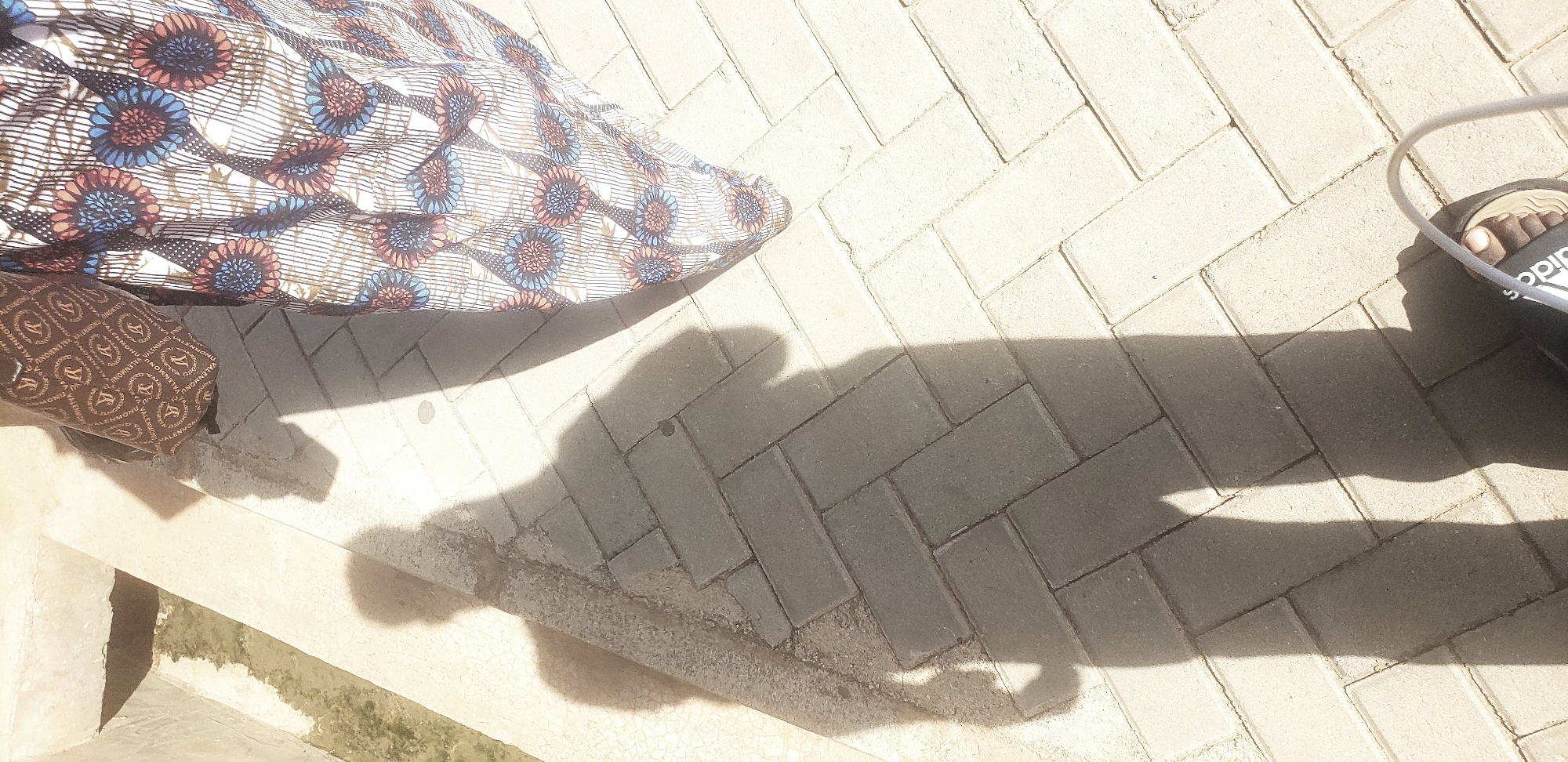 The image depicts an outdoor paved area, identified as being in Narayi, Nigeria, though no specific geographical markers are discernible within the frame to confirm this location. The scene is illuminated by strong direct sunlight, casting prominent shadows.
In the upper right portion of the image, a person is partially visible, primarily their torso and lower body, clad in a patterned garment. The upper part of the garment, specifically a sleeve, is brown with a repeating pattern that includes the text "VALENMONRO" and a stylized 'V' logo. The lower part of the garment, extending downwards, is predominantly white with a distinct pattern of blue and reddish-brown circular floral motifs and vertical dark lines.
The ground is paved with light-colored rectangular interlocking bricks, laid in a herringbone pattern in the central and right areas, and a running bond pattern towards the left. On the far left, a raised concrete ledge borders a narrow, shallow concrete channel or ditch, which contains some dark, possibly organic, material or stagnant water.
Two distinct human shadows are cast on the paved surface. The most prominent shadow, occupying the lower-left to mid-right sections, is that of a person, likely the individual taking the photograph. This shadow clearly outlines a human torso and legs. A smaller, fainter shadow of a head and upper body is visible near the concrete ledge in the upper-left, partially overlapping with the main shadow. In the bottom right corner, a human foot with dark skin is visible, wearing a black slide sandal with "adidas" imprinted in white on the strap. A light-colored, thin, curved object, possibly a cable or strap, extends from the right edge of the frame near the sandal.