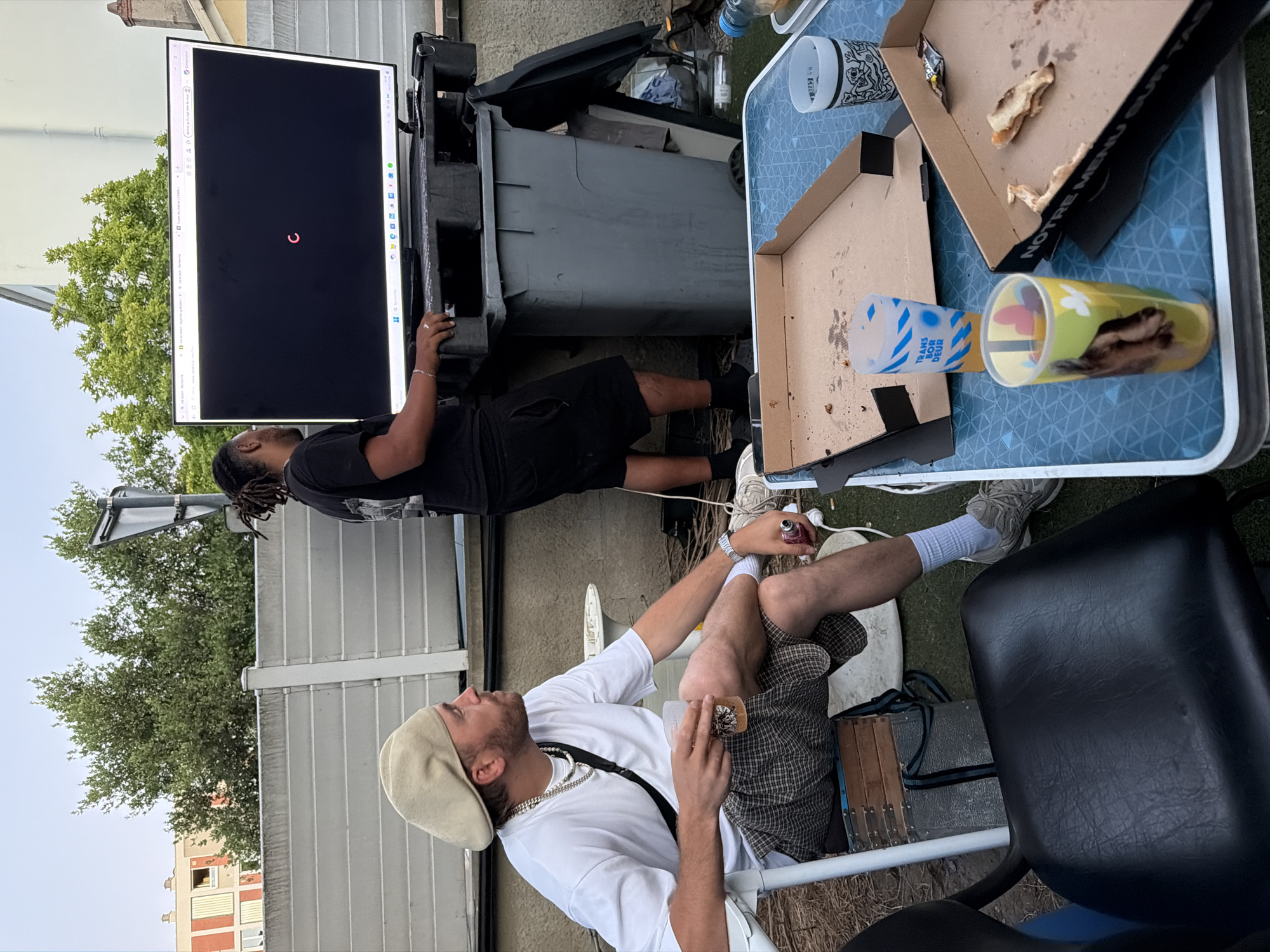 The image shows an outdoor scene in Villeurbanne, France, likely a backyard or terrace. Two men are present. One man, wearing a beret, white t-shirt, and checkered shorts, is seated in a white chair, holding a clear plastic cup. The other man, with dreadlocks and wearing a black t-shirt and shorts, stands in front of a large television screen which displays a loading symbol. The TV is placed on a black pallet atop a grey dumpster. A blue and white patterned table is set up in the foreground, with pizza boxes and plastic cups on it. The background shows trees, a fence, and apartment buildings, consistent with an urban environment. The lighting suggests it is late afternoon or early evening.