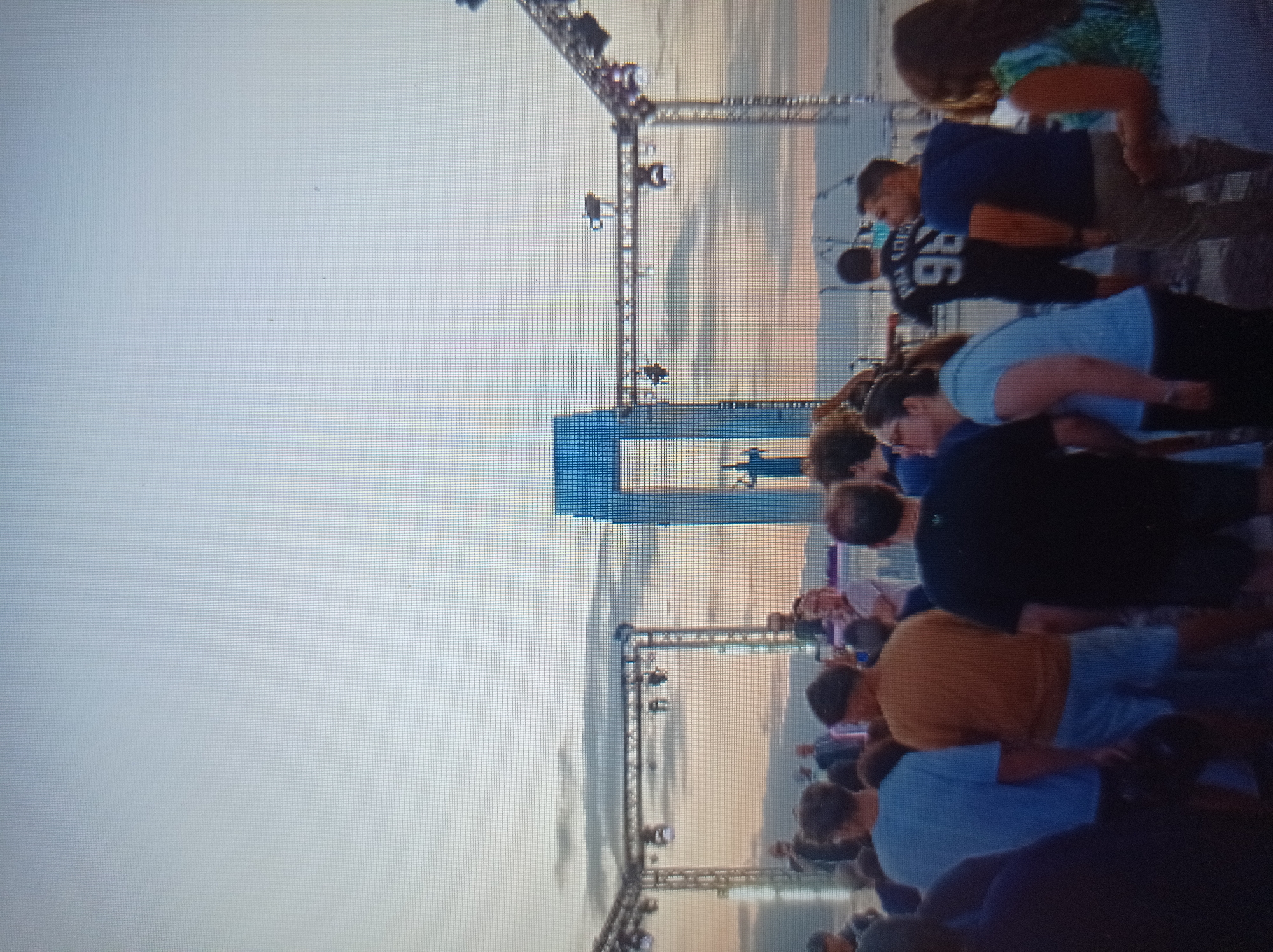The image depicts an outdoor gathering with a crowd of people observing a stage setup. In the background, a large, blue, arch-like structure stands prominently, with a silhouette of a person in front of it. The sky is a gradient of light blue and grey, suggesting either dawn or dusk, with some clouds visible. There are truss structures with spotlights positioned around the stage area. The foreground is occupied by a crowd of individuals, some facing the stage and others with their backs to the camera. The overall scene appears to be an event or concert taking place in an open area. The prompt states this image is in the city of Mukachevo, Ukraine, however, there are no visual cues in the image that confirm this specific location.