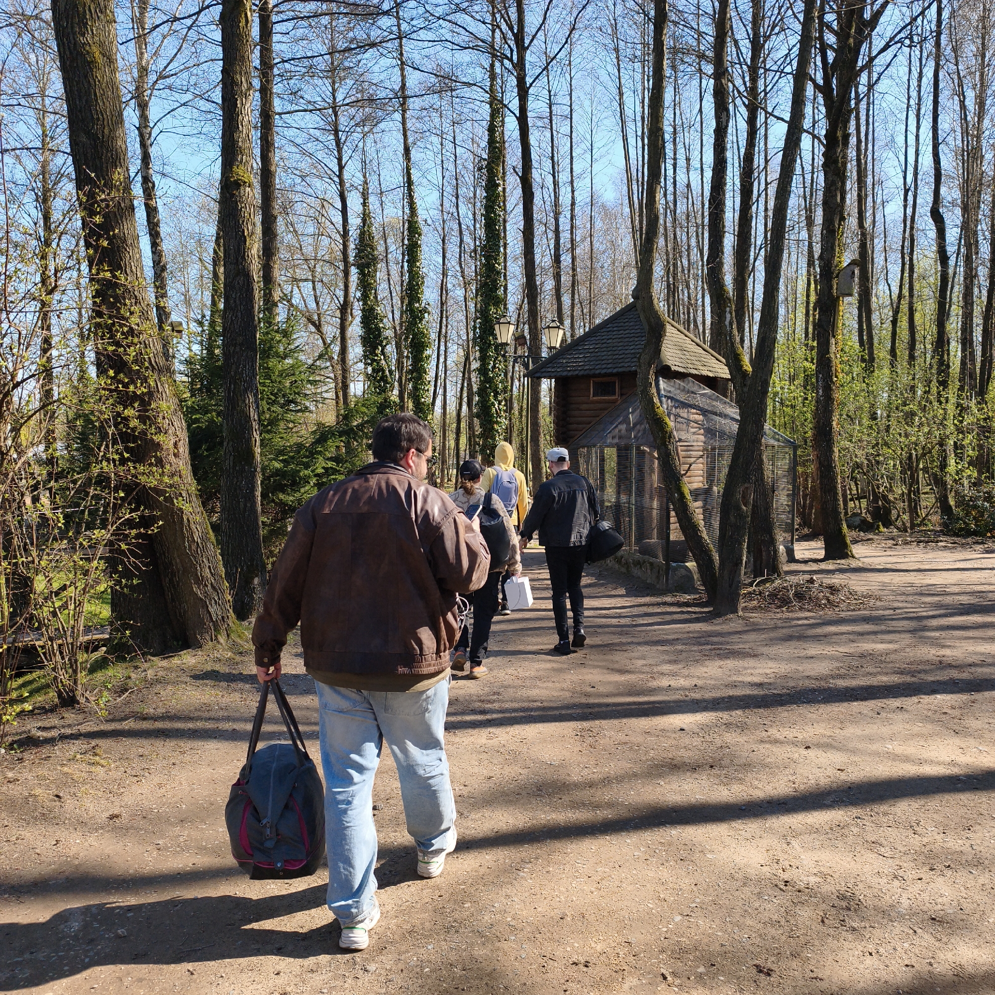 A group of people walks along a sunlit dirt path through a wooded area. In the foreground, a man, seen from behind, wears a brown leather jacket, light blue jeans, and white sneakers, carrying a dark gray duffel bag with pink accents. Behind him, other individuals are visible, including one in a yellow hoodie with a backpack and another in a black cap and jacket carrying a black bag.
The path is surrounded by numerous tall, slender trees with mostly bare branches, suggesting early spring as some smaller trees and bushes show new green buds and leaves. Patches of green moss cling to the trunks of larger trees. The sky above is clear and bright blue. To the right of the path stands a rustic wooden structure with a dark, tiled roof and mesh-covered sides, resembling a large aviary or animal enclosure. Two vintage-style street lamps are also visible near this structure. Long shadows are cast across the dusty path by the trees and people, indicating bright sunshine, likely during midday or early afternoon. A small birdhouse or nesting box is perched on one of the tree trunks in the background. No visible text is present in the image.