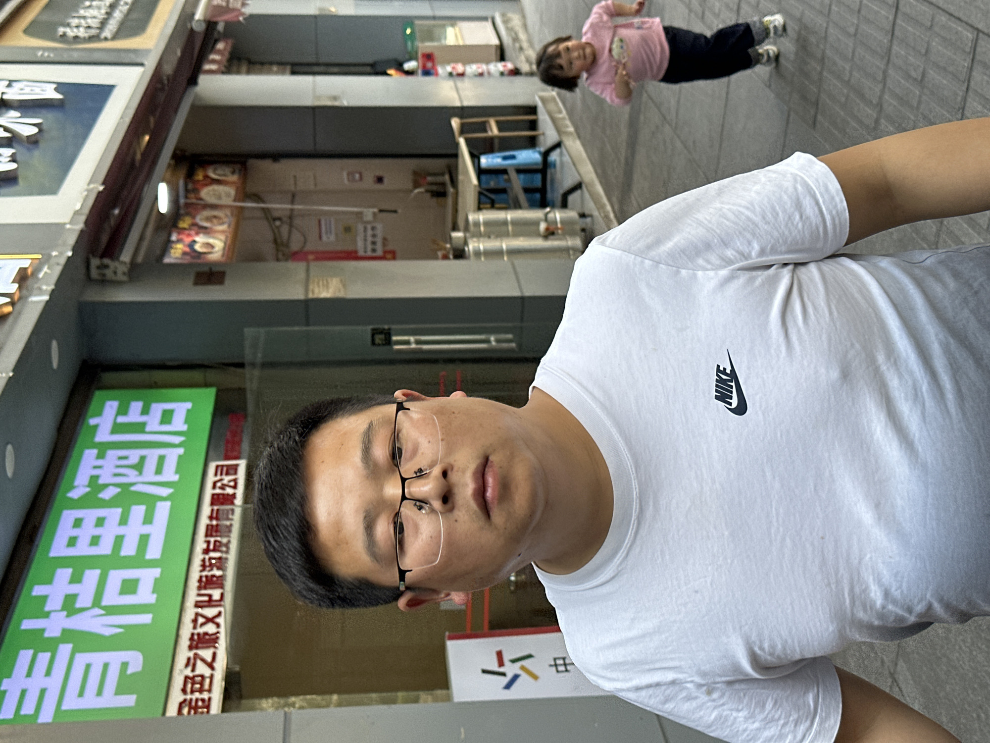 A man wearing glasses and a white Nike t-shirt stands on a sidewalk in the Yuzhong District of China. In the background, a child in a pink shirt and dark pants stands further down the sidewalk. A sign with Chinese characters is visible to the left, and various structures and signage are further in the background.
