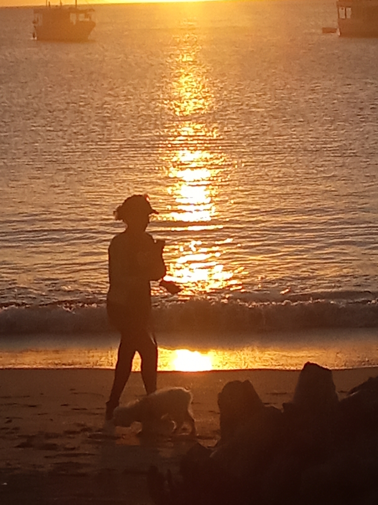 The image displays an outdoor scene captured during the golden hour, likely sunset, on a beach in Juan Griego, Venezuela. A person, rendered in dark silhouette, walks from left to right near the water's edge, wearing what appears to be a cap or has hair tied up, a shirt, and pants. Their head is oriented towards the right. Accompanying the person is a small, light-colored animal, possibly a dog, walking close to their feet, also moving rightward. The ocean or bay occupies the midground, with strong, shimmering golden sunlight reflecting directly on its surface from the low-angle sun at the horizon. Small waves are visible breaking onto the sandy shore in the immediate foreground. In the distant water, two boats are silhouetted against the bright horizon; one is positioned on the far left, and another is on the far right. The immediate foreground on the right contains several dark, irregularly shaped objects, consistent with rocks or debris. The photograph's dominant colors are warm oranges, golds, and dark browns, with subjects appearing as dark silhouettes against the luminous background. The image has a soft focus.