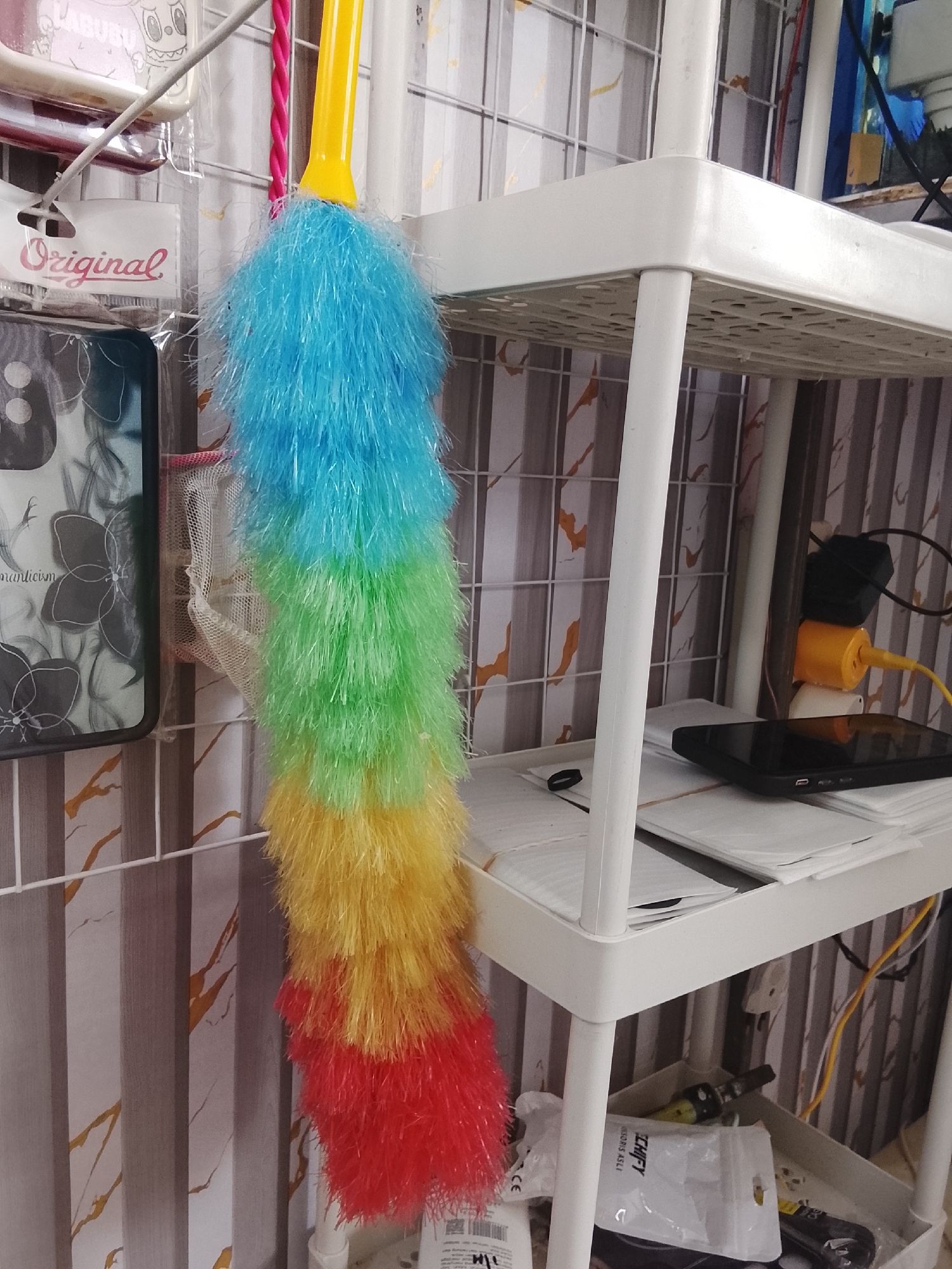 An indoor scene in Mee, Indonesia shows a white plastic shelf unit with various items on it and hanging from it. A yellow-handled duster with brightly colored, feathery strands hangs vertically from the left side of the scene. The duster has segments of blue, green, yellow, and red. To the left of the duster, a black smartphone case with a floral design is attached to a wire rack. A white plastic tag with the word "Original" in red script is visible below the smartphone case. On the shelves of the unit, there are stacks of white envelopes, a black smartphone, and electrical adapters plugged into a yellow outlet. The background features vertical striped wallpaper with orange accents.