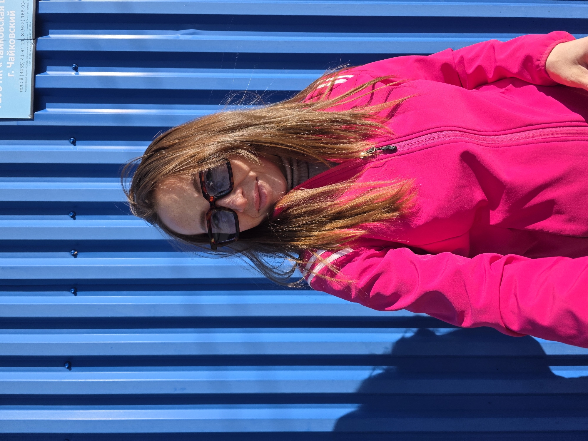 A woman with long brown hair and sunglasses is lying on a blue corrugated metal surface. She is wearing a bright pink jacket with white stripes on the sleeves. The background is a blue corrugated metal fence or wall. A sign is partially visible on the left side of the image, with text in Russian mentioning the city of Chaikovsky and phone numbers.