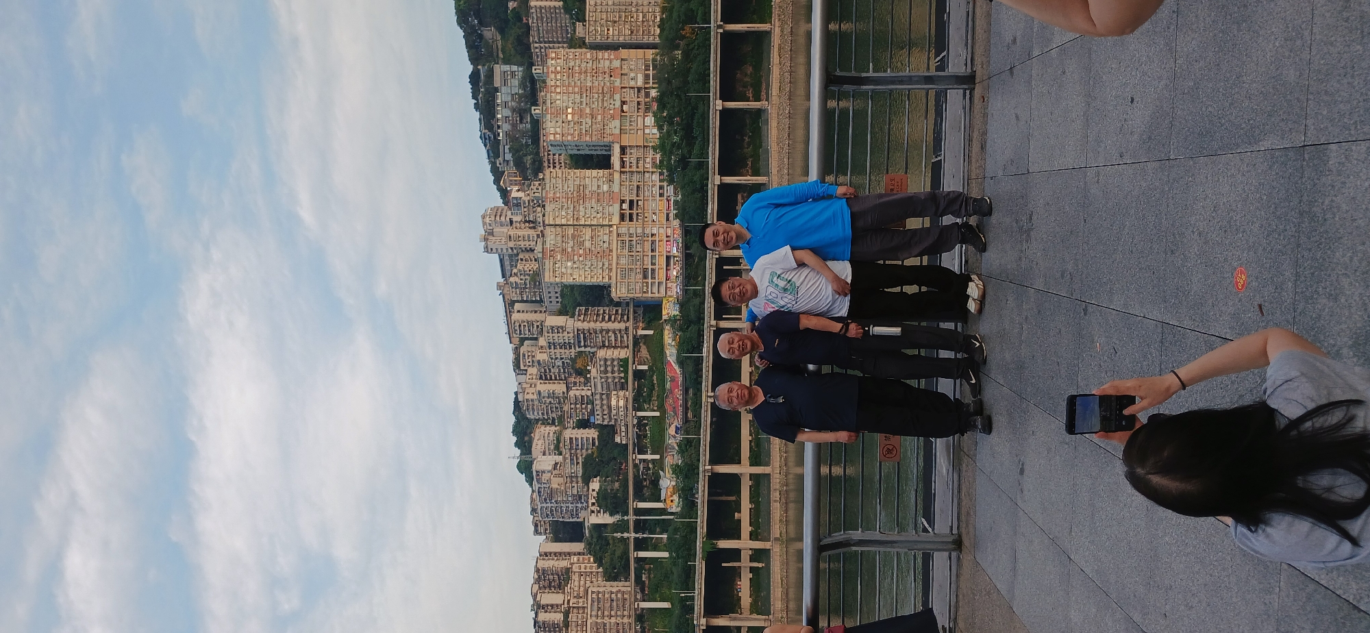 Four men stand in a line along a gray sidewalk, facing the camera. A woman with dark hair and a gray shirt holds a phone, taking a photo. Behind the men, there's a metal railing, a river, and a dense urban cityscape of tall buildings on a hillside. The sky is light blue with some clouds. The location is in the Yuzhong District of China.