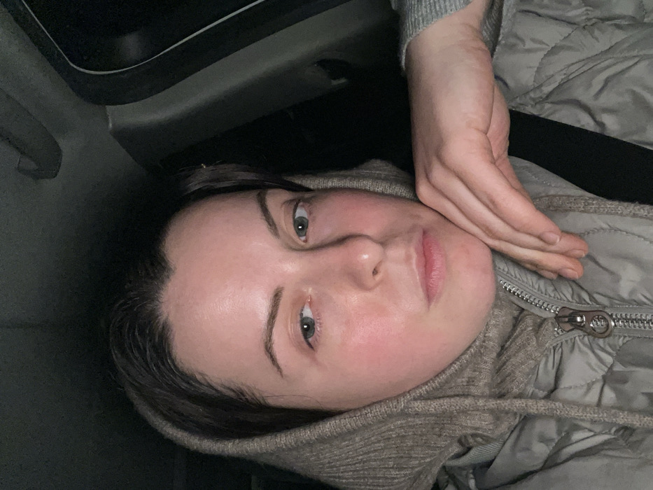 A woman lies on her side, seemingly in a car, her head resting on her hand. She has fair skin, light eyes, and brown hair. A gray puffer jacket is partially visible, with a zipped closure near her neck. The background appears dark, possibly the interior of a vehicle. Her face is angled towards the camera, and her expression is neutral.