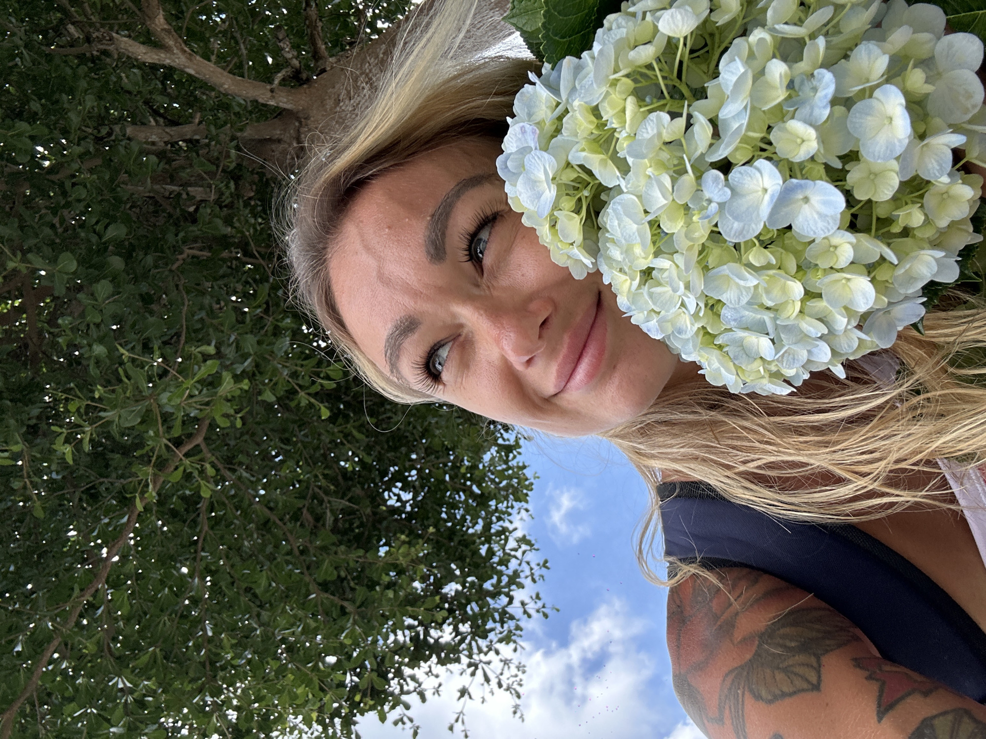 A woman with blonde hair and tattoos on her arm smiles as she looks up at the sky, holding a bouquet of light blue and white hydrangeas. She is positioned beneath a large tree with green foliage, and the sky above is a clear blue with a few wisps of clouds. The scene suggests an outdoor setting, possibly a park or garden. This image does not contain any discernible landmarks or elements that would place it specifically within Da Lat, Vietnam.