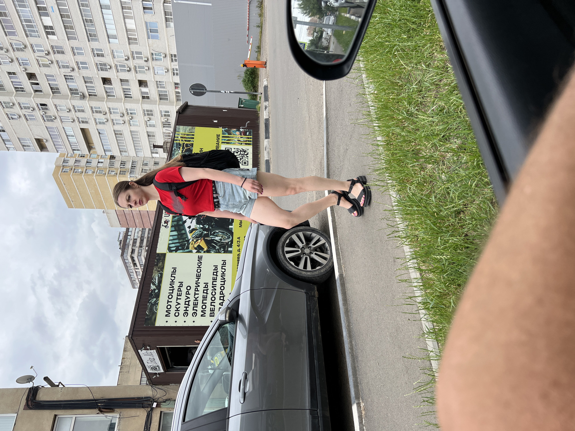 A young woman with a ponytail, wearing a red t-shirt, denim shorts, and black platform sandals, walks along a paved sidewalk. She is carrying a black backpack. To her left is a gray car. Behind her are buildings, including a multi-story apartment complex. A sign with Russian text and images of motorcycles is visible on a building to her left. The scene appears to be in Krasnodar, Russia, based on the context provided in the prompt.