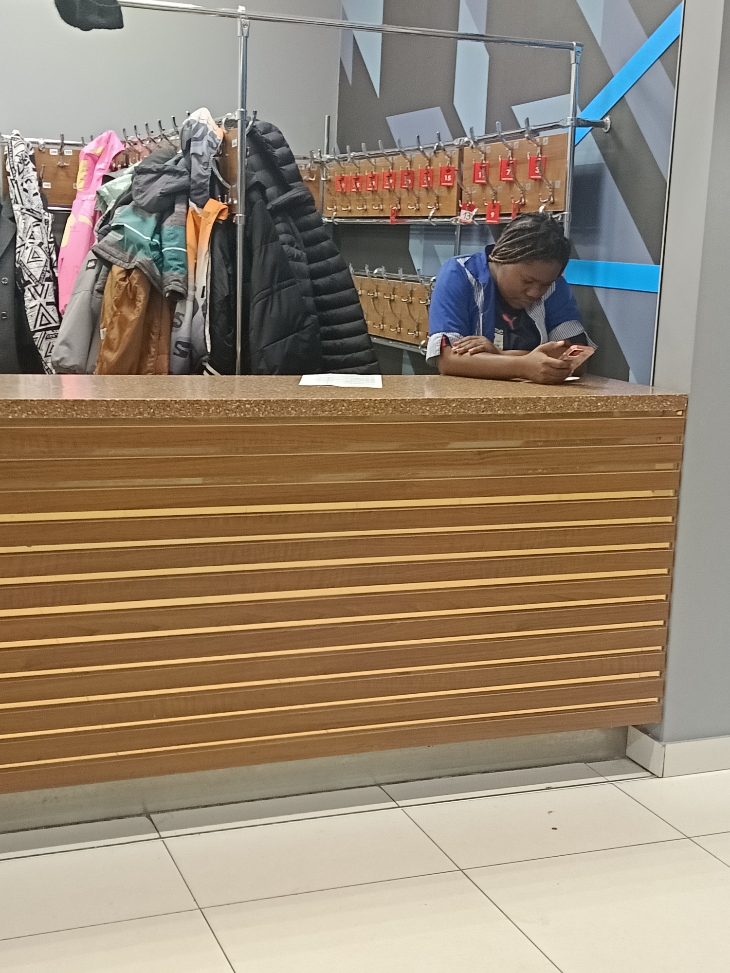 The image shows an individual with braided hair, wearing a blue and white striped shirt, looking at a mobile phone behind a wood-paneled counter. Behind the counter is a rack of hanging coats, and another rack holding numbered keys. The location may be a coat check area, potentially in Izhevsk, Russia.