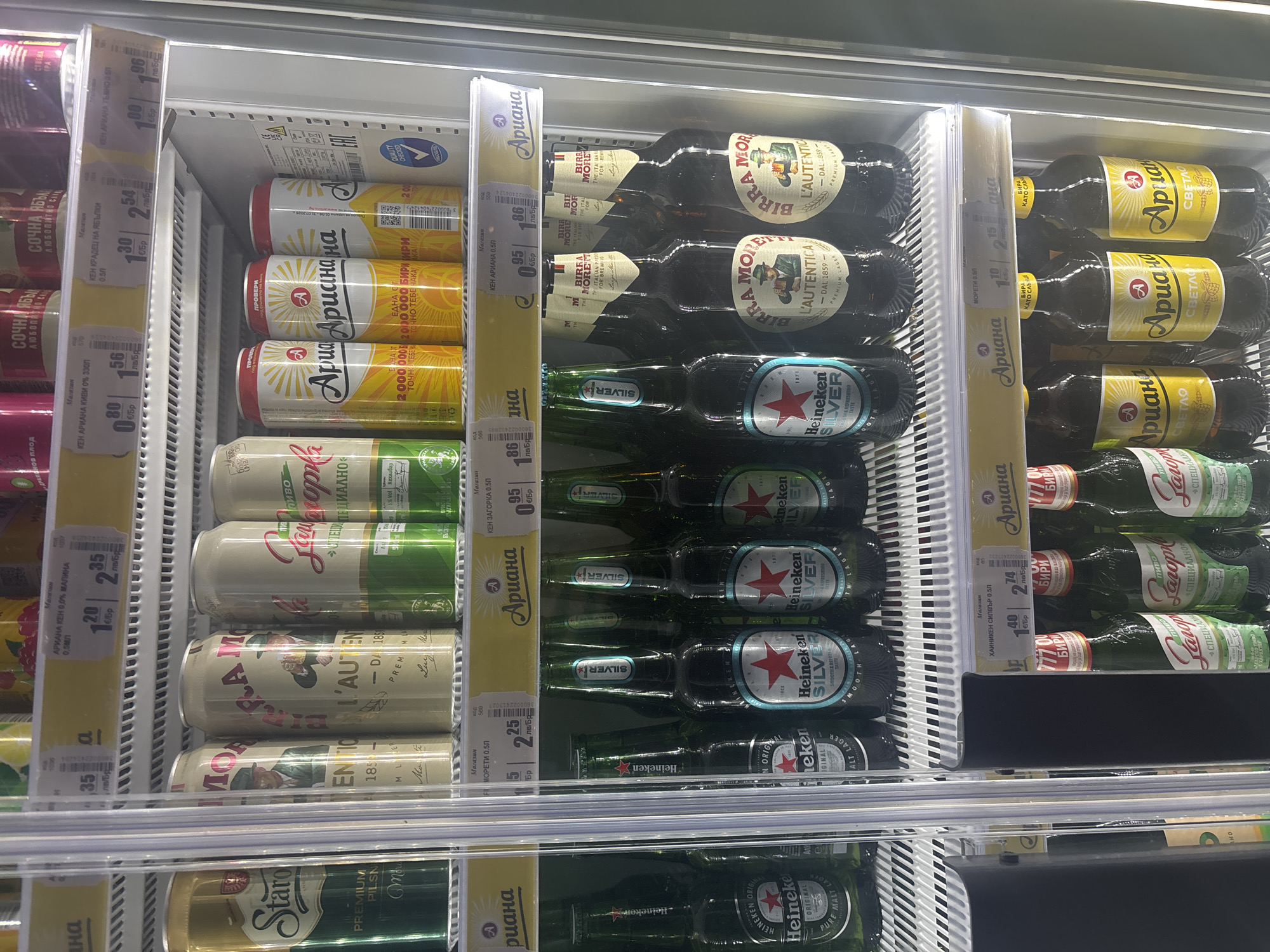 The image depicts a refrigerator shelf in a store, likely in Plovdiv, Bulgaria, given the Bulgarian Cyrillic text on the labels. The shelf contains various brands of beer, including cans and bottles. The labels are mostly in Bulgarian, with some English. Beer brands include "Ariana," "Zagorka," "Birra Moretti," and "Heineken Silver." Pricing information is displayed on shelf edge labels, indicating the price per unit in local currency.
