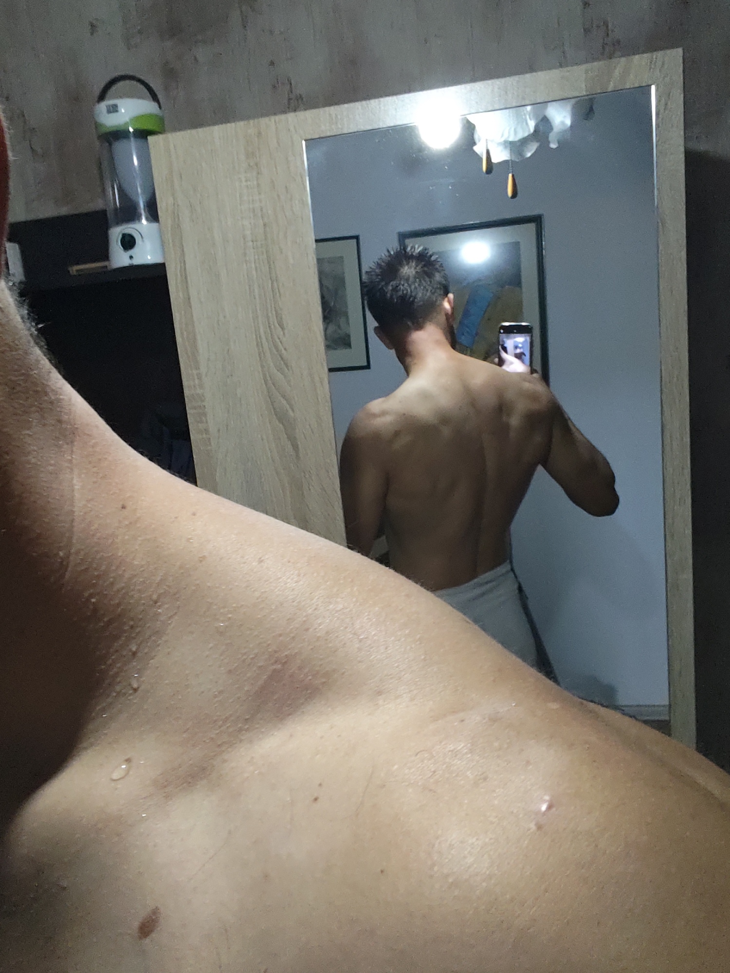 A shirtless man is taking a selfie in front of a mirror. The man is facing away from the viewer and holding a phone in his right hand. The mirror is reflecting his back and the surrounding room. A framed picture is on the wall behind him. A lamp is on a shelf next to the mirror. The man appears to have recently showered, as there are water droplets on his skin. The location is unknown.