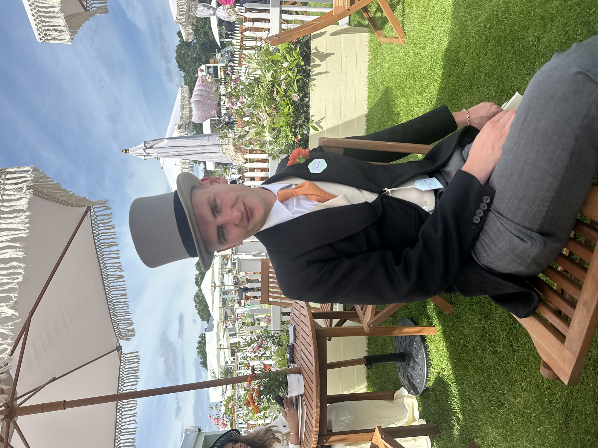 A man in formal attire, including a black tailcoat, grey trousers, and a grey top hat, sits on a wooden chair in a grassy outdoor area. He is looking towards the camera with a slight smile. He is wearing an orange tie and a corsage on his lapel. Behind him, there are other people, white umbrellas with fringe, and decorative floral arrangements. In the background, there is a white fence and trees. The scene appears to be set at Ascot Racecourse in Ascot, United Kingdom, likely during an event with a formal dress code.