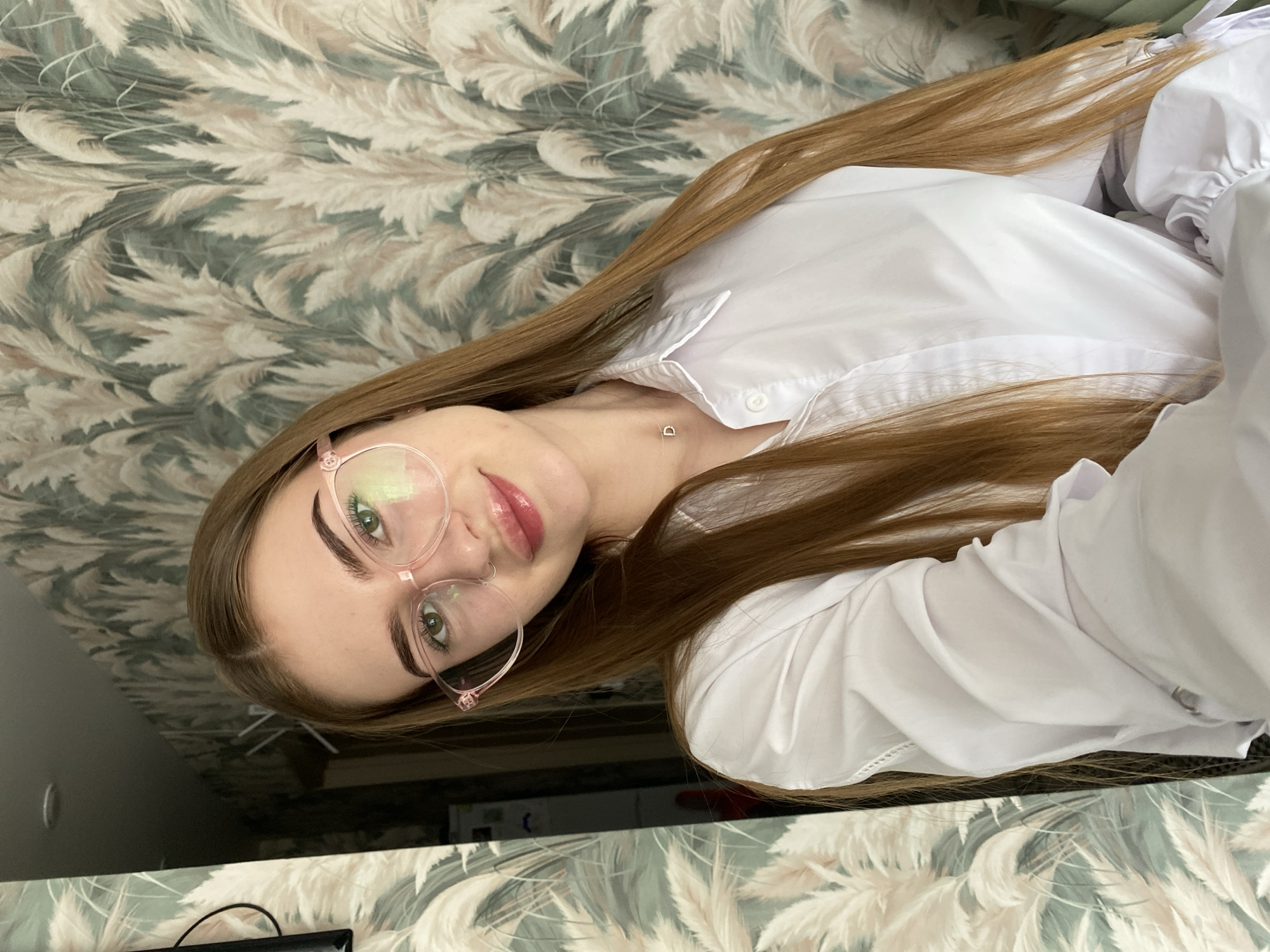 A young woman with long, brown hair and glasses is taking a selfie in front of a wall with a floral wallpaper. She is wearing a white collared shirt. The background wallpaper appears to be a pale green with a pattern of tall, feathery reeds in shades of white and pink. The room is dimly lit, and there is a door frame visible to the left of the woman.