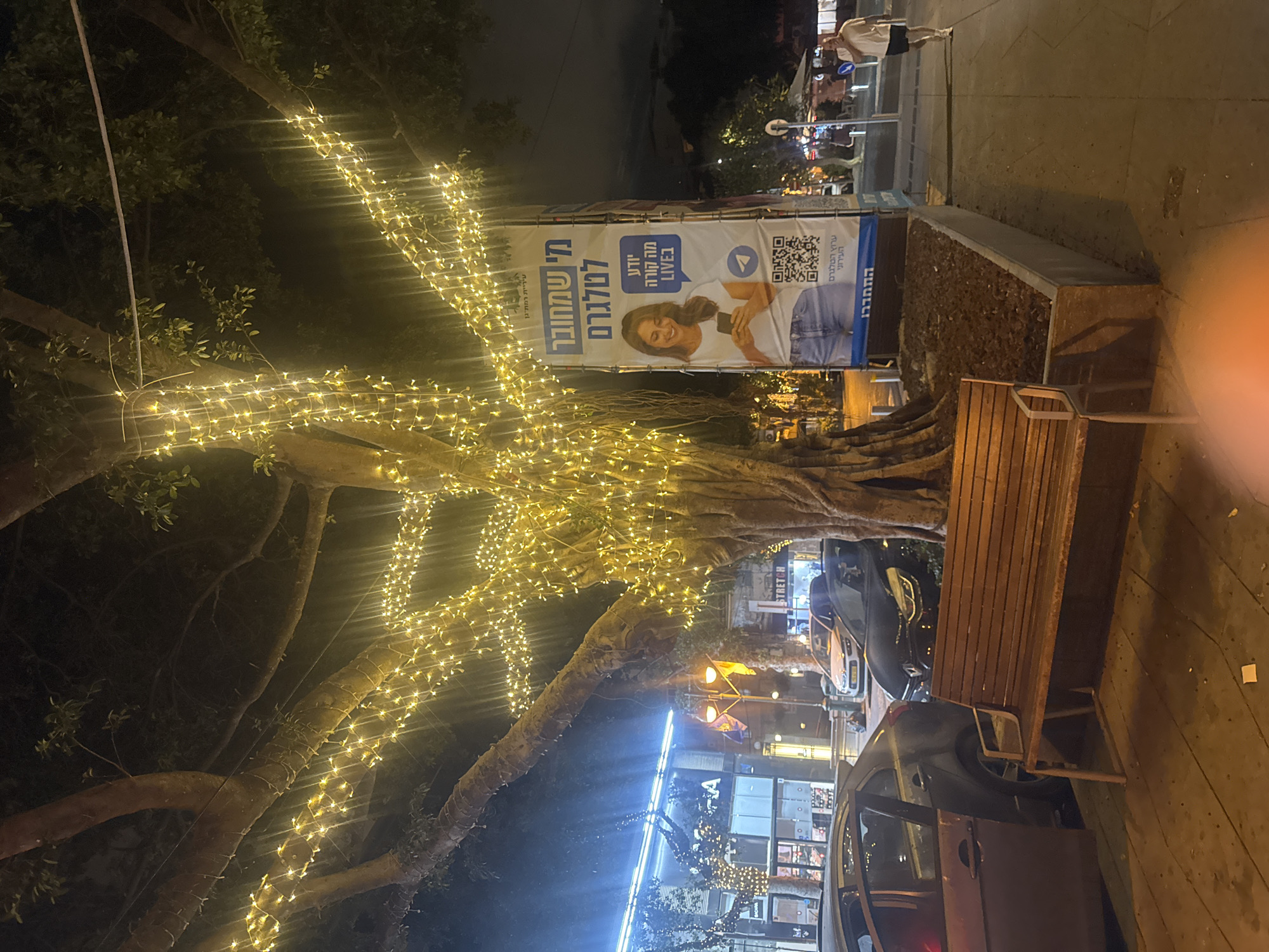 Here is a detailed description of the image:
The image captures a nighttime scene in what appears to be a commercial area of Ramat Gan, Israel. An advertising banner is visible, advertising a "LIVE" Telegram channel with Hebrew text.
The scene is partially illuminated with strings of yellow lights wrapped around a large tree. A wooden bench sits beside a tree planter. In the background, the image captures a glimpse of a street with buildings, signs (including one labeled "STRETCH"), and parked cars. There are people walking in the background as well.