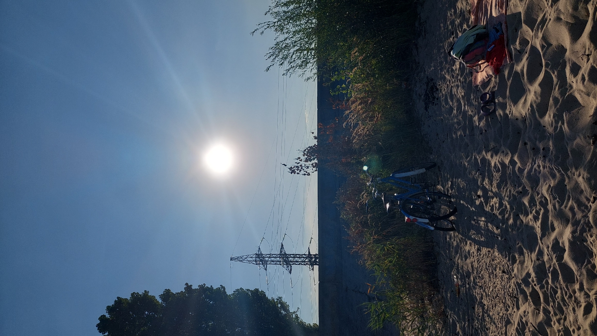 The image depicts a sunny beach scene in Kozarovychi, Ukraine, likely along the Kyiv Reservoir, given the presence of high-voltage power lines and a pylon stretching across the horizon. In the foreground, light brown sand is textured with ripples and shadows cast by the low-lying sun, which is positioned high in the bright blue sky, creating a strong glare. Two blue bicycles are parked on the sand, leaning against a thicket of dry grass and green reeds that separate the beach from a concrete barrier. To the right, a red towel is spread on the sand, with a backpack and a pair of dark sandals resting on it. A large tree with dense green foliage is visible on the far left, silhouetted against the sky, and a power pylon stands prominently behind the concrete barrier. The overall atmosphere is one of serene, late afternoon or early morning outdoor recreation.