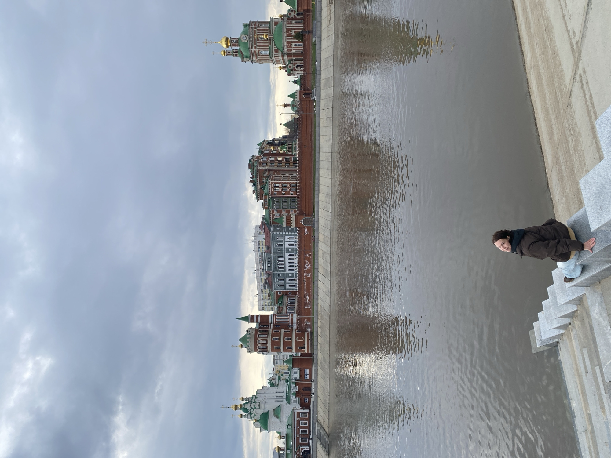 The image, when correctly oriented, depicts a picturesque urban waterfront scene in Yoshkar-Ola, Russia. A woman in a dark brown jacket and light-colored pants is seated on a gray stone staircase, with her feet near the water's edge on the right side of the frame. She is looking towards the viewer with a slight smile.
Across a relatively wide river, a vibrant skyline of unique architecture stretches along the far bank. The buildings feature predominantly red brick facades, often adorned with white trim, green roofs, and numerous golden onion domes and crosses characteristic of Russian Orthodox churches and cathedrals. Some structures also show elements resembling European or fairytale-like architecture, with varied window shapes and decorative details. The buildings are a mix of residential or commercial structures interspersed with church-like edifices, creating a distinctive and colorful panorama.
The river itself is a muted brownish-gray, reflecting the cloudy sky and showing ripples that catch what little light is available. The sky above is overcast with soft gray clouds, suggesting a cloudy day or perhaps late afternoon without direct sunlight. The overall atmosphere is calm and serene. There is no visible text in the image.