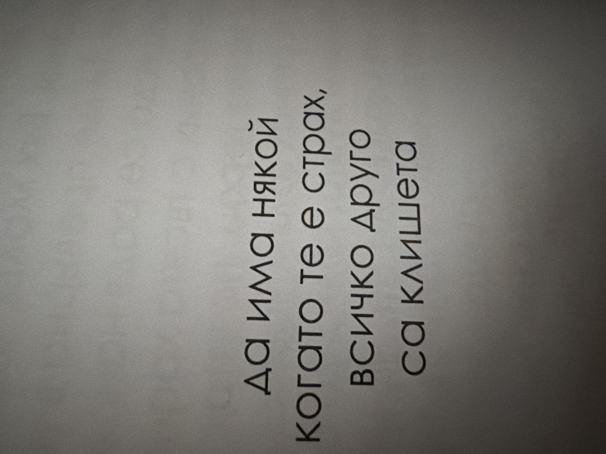 The image contains Bulgarian text printed on a textured, light gray background. The text is centered on the page and appears to be a quote or saying. The first line reads "да има някой" which translates to "to have someone." The second line reads "когато те е страх," meaning "when you are afraid." The third line states "всичко друго" translating to "everything else." The final line reads "са клишета" which means "are clichés." The text is written in a sans-serif font and is black in color. There are no people or other objects visible in the image. The background has a subtle gradient, appearing slightly darker towards the bottom right. There is no information available to indicate the location in the city of Pazardzhik, Bulgaria.