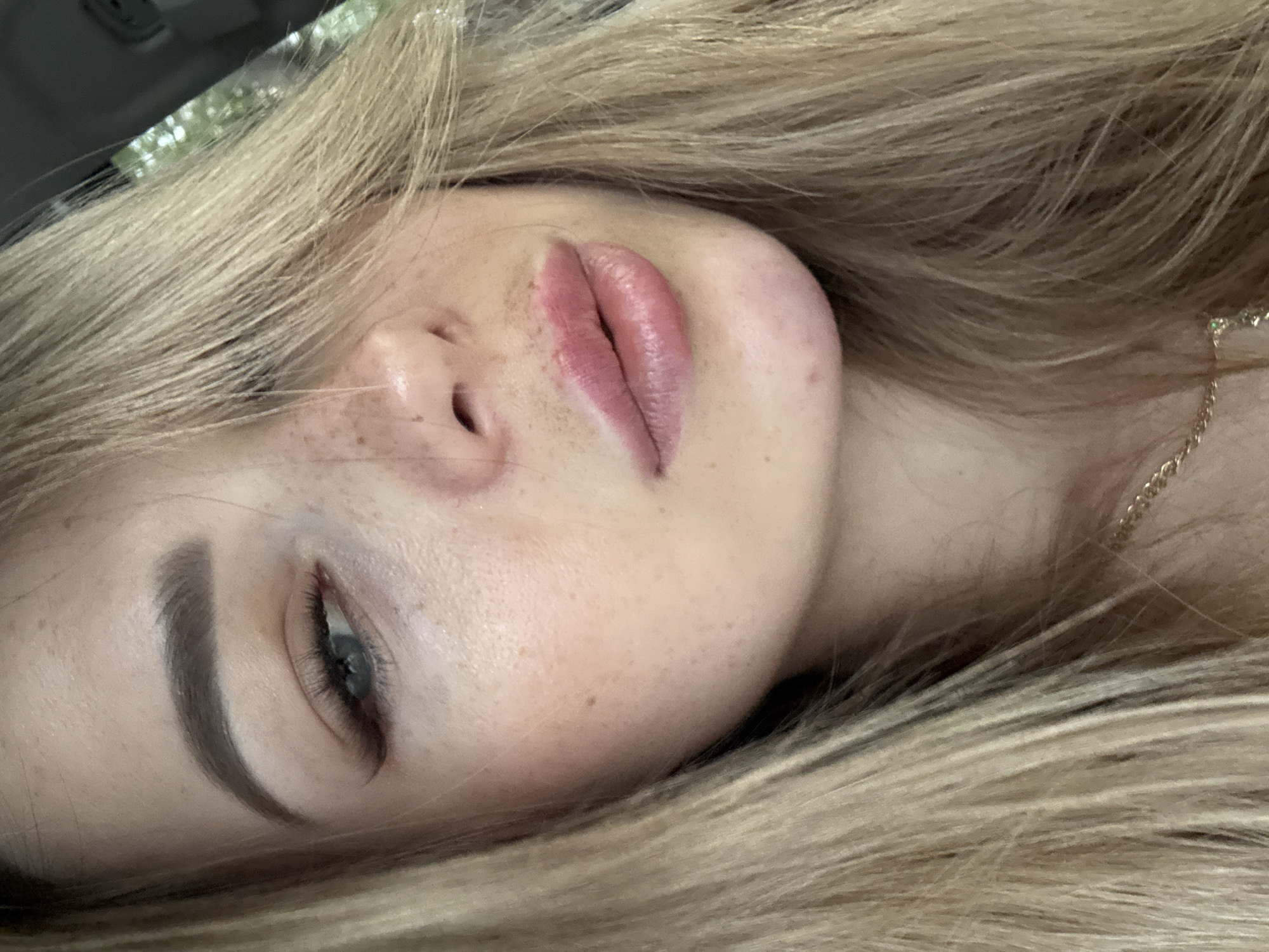 A close-up, portrait-style photograph captures the left side of a young woman's face. She has blonde hair that partially obscures her left eye and cheek. Her skin is fair with visible freckles across her nose and cheeks. She is wearing subtle makeup, including eyeliner and lipstick. Her lips are full and painted a pinkish-nude shade. A thin gold chain necklace is visible around her neck. The background is blurred, but appears to be outdoors with hints of greenery and possibly the interior of a vehicle. The lighting suggests it is daytime. The image is shot from a slightly upward angle, focusing on her facial features.