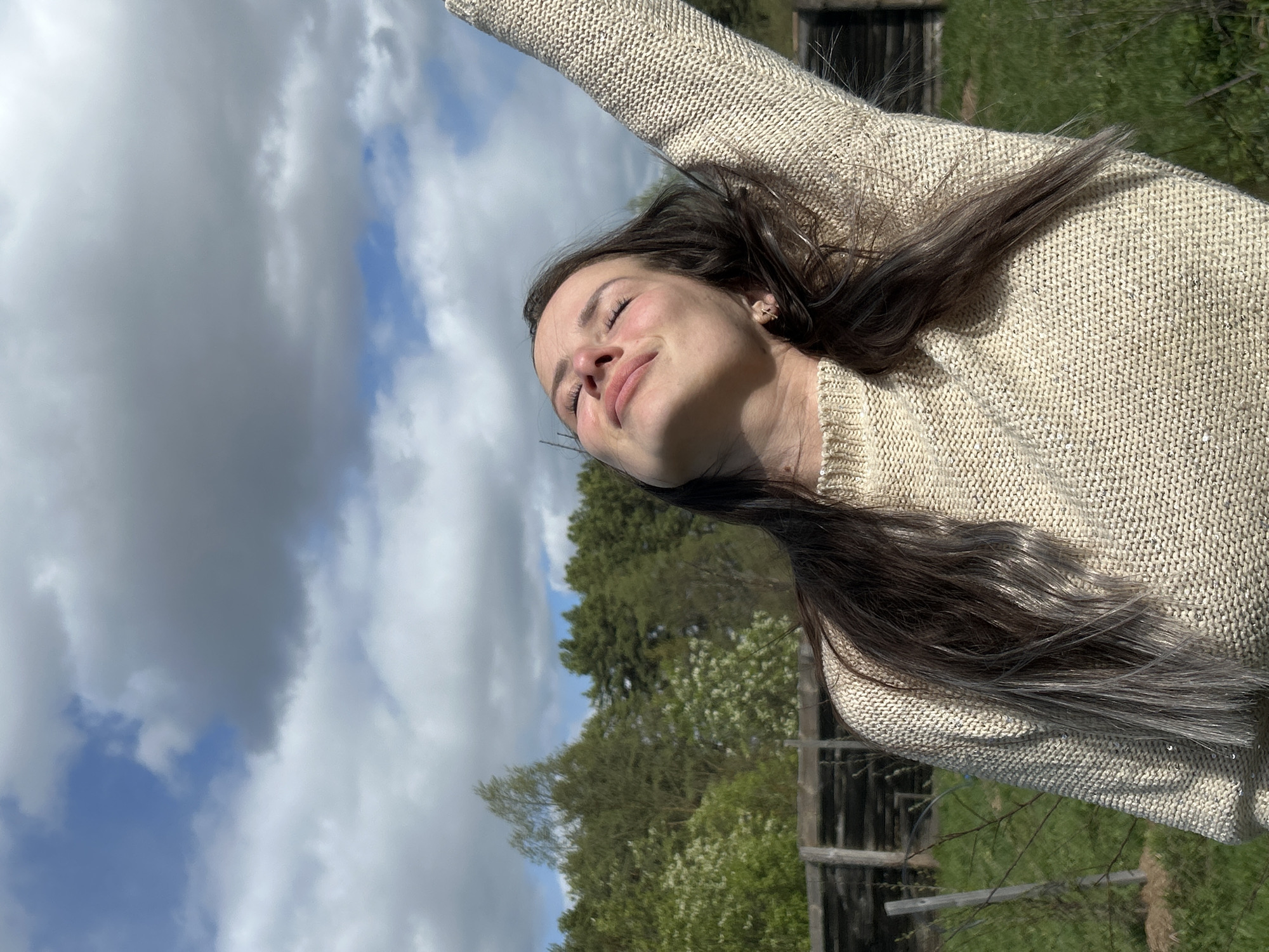 A young woman with long brown hair stands outdoors, facing the sky with her eyes closed. She is wearing a beige knitted sweater and has her right arm raised. The background features a sky with cumulus clouds, trees with green foliage, and a weathered wooden structure. The location is likely Rusalevka, Russia.